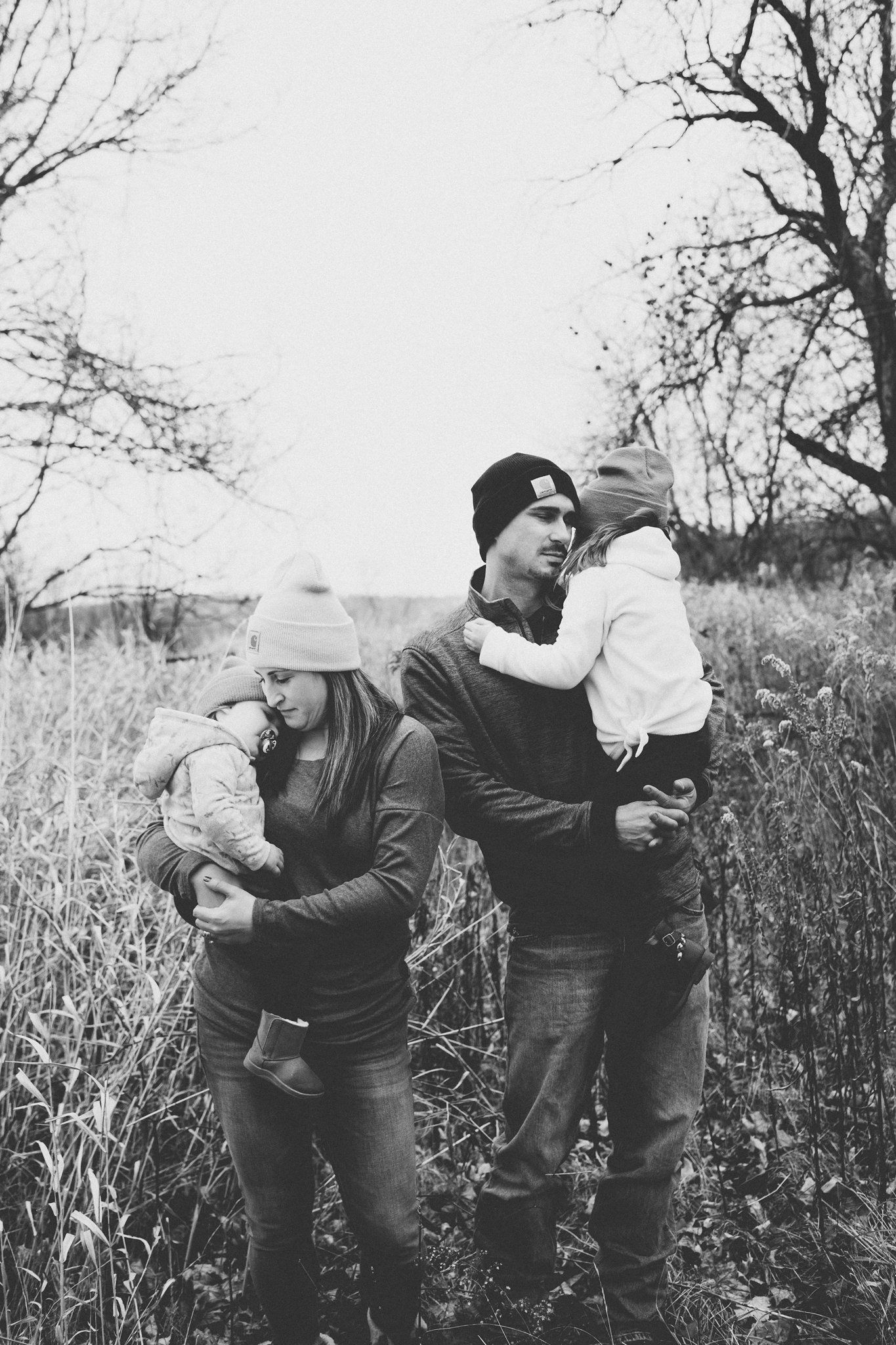 A couple holds their young children while standing together in a rustic outdoor field setting.