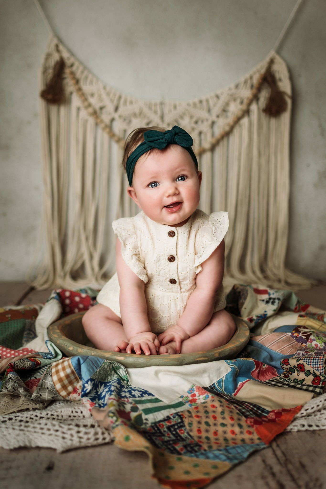 Baby wearing cream colored romper and teal headband sits on vintage quilt with macrame backdrop.
