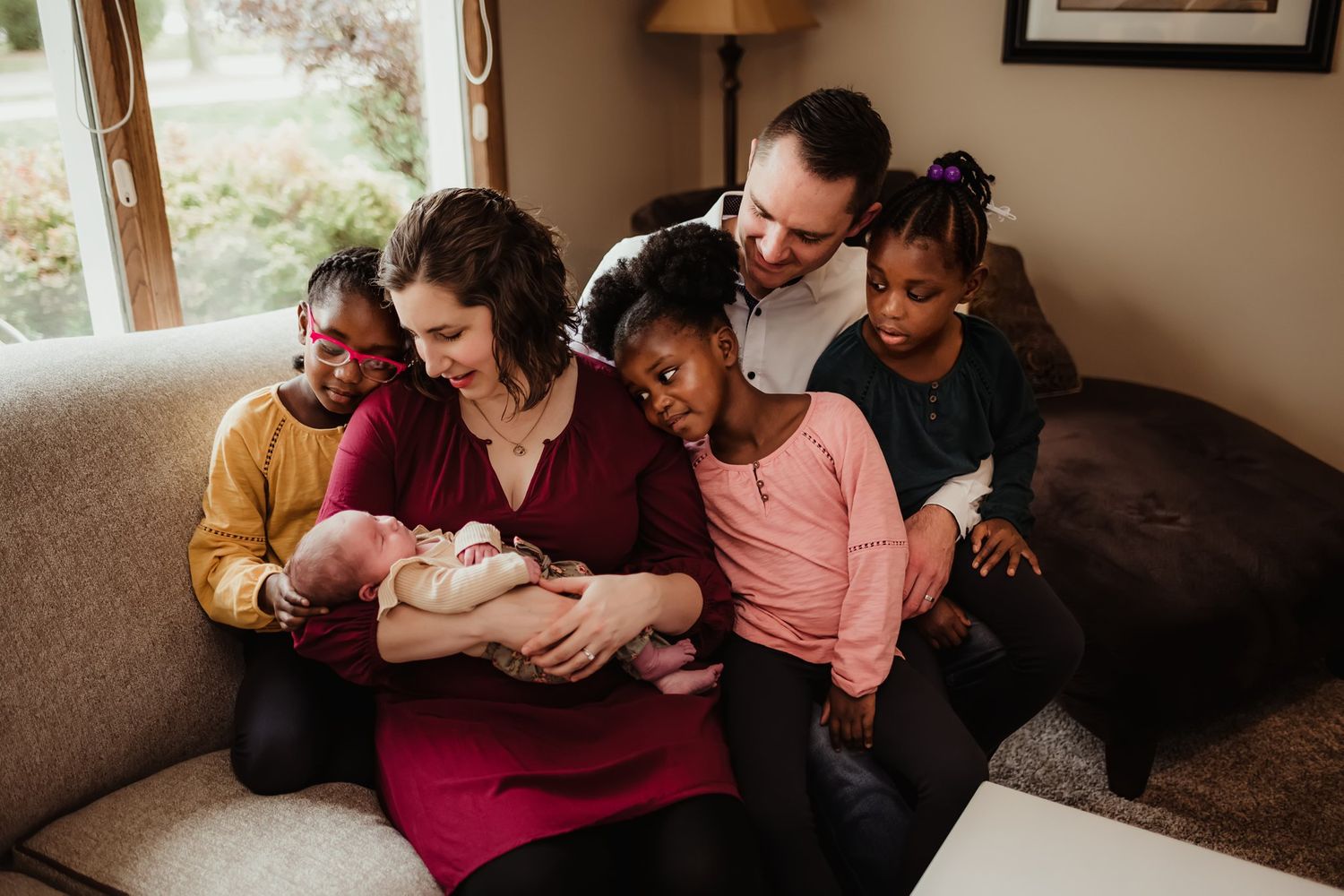 A family gathers closely together on a beige couch in a warm-lit living room with a newborn baby.