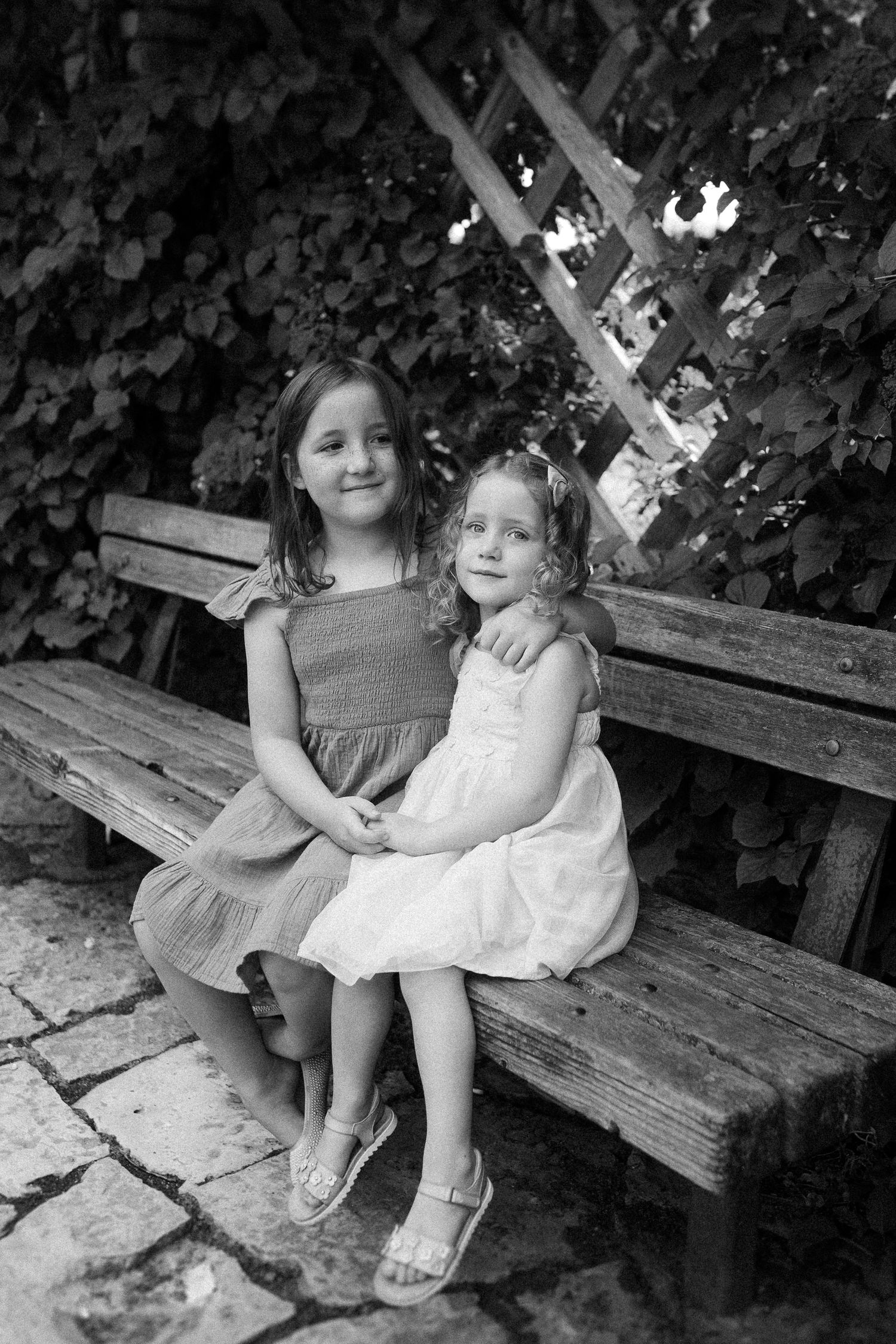 Black and white portrait of two children sitting together on a wooden bench in a garden setting.