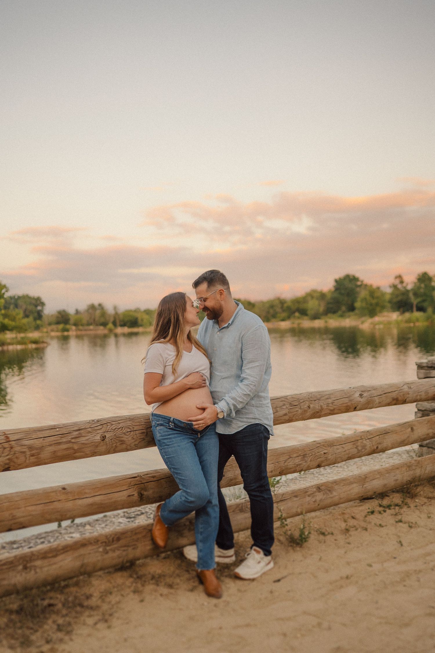 Couple shares romantic sunset moment by lake with wooden fence during golden hour photography session.