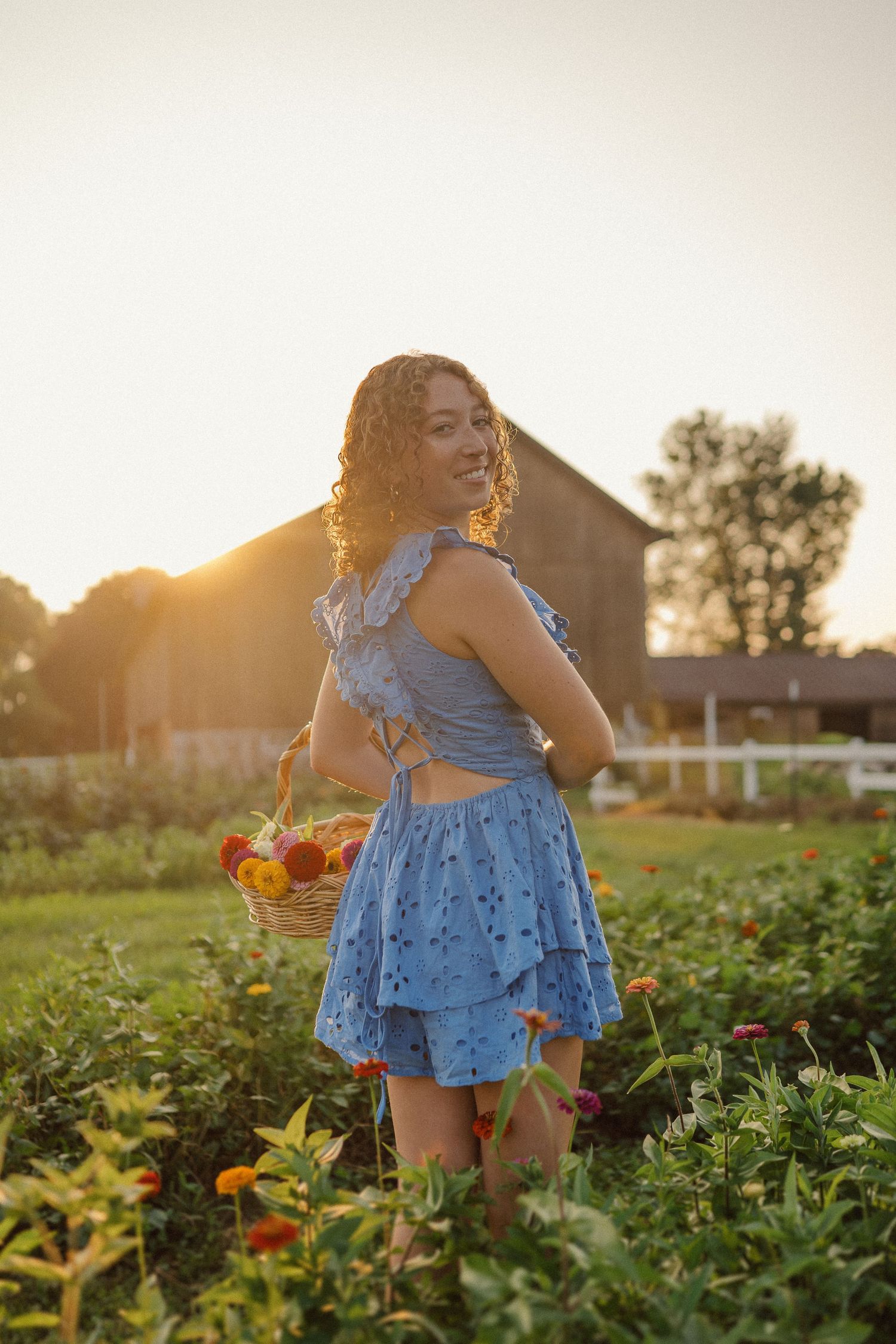 Person in a blue summer dress stands in a garden holding a basket during golden hour sunset with a barn in background.