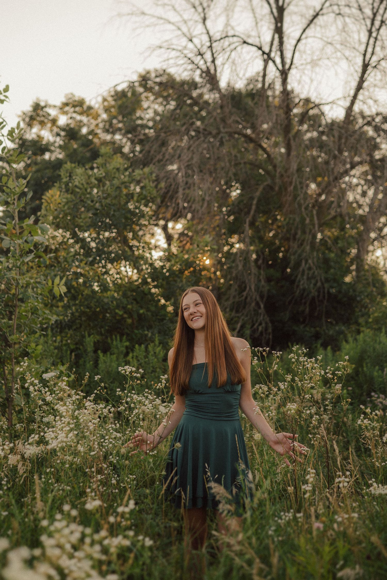 Person in dark green dress stands joyfully among wildflowers during sunset.