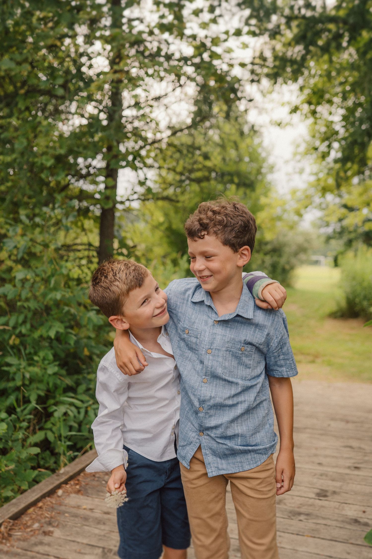 Two children share a heartwarming moment while walking together on a wooden boardwalk surrounded by lush greenery.