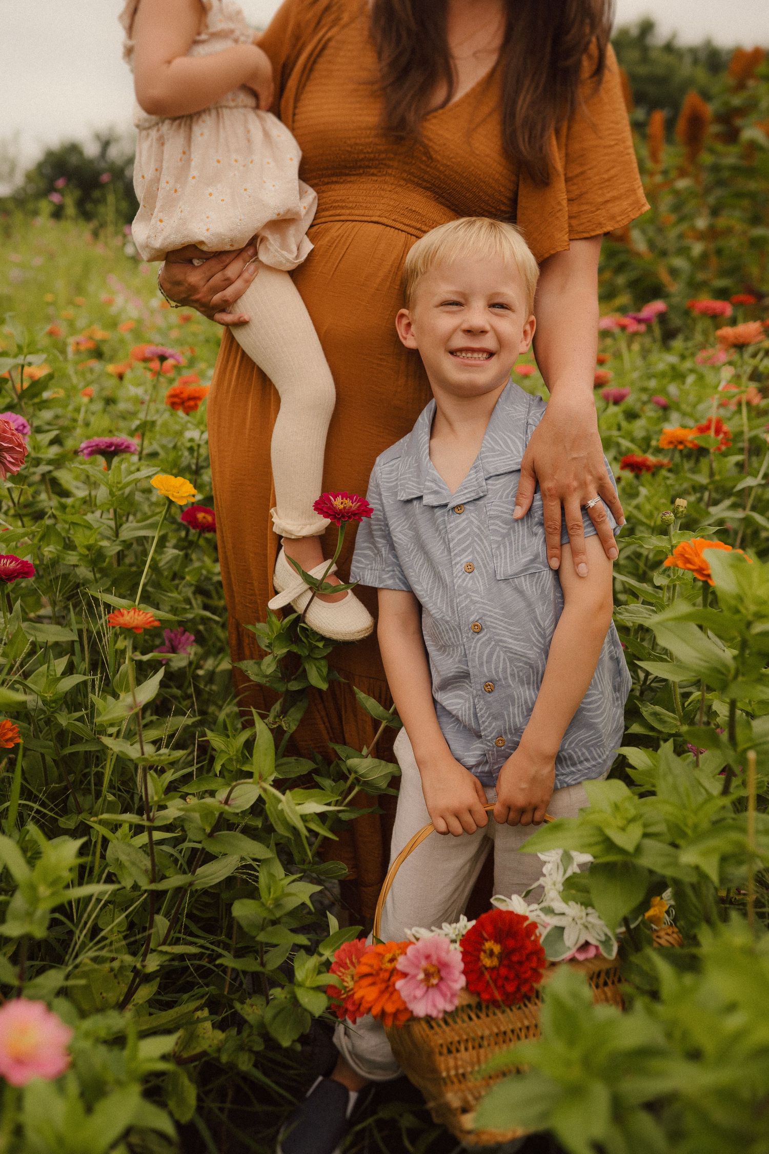 In a blooming garden of zinnias, someone in an orange dress stands with a small child wearing blue overalls.