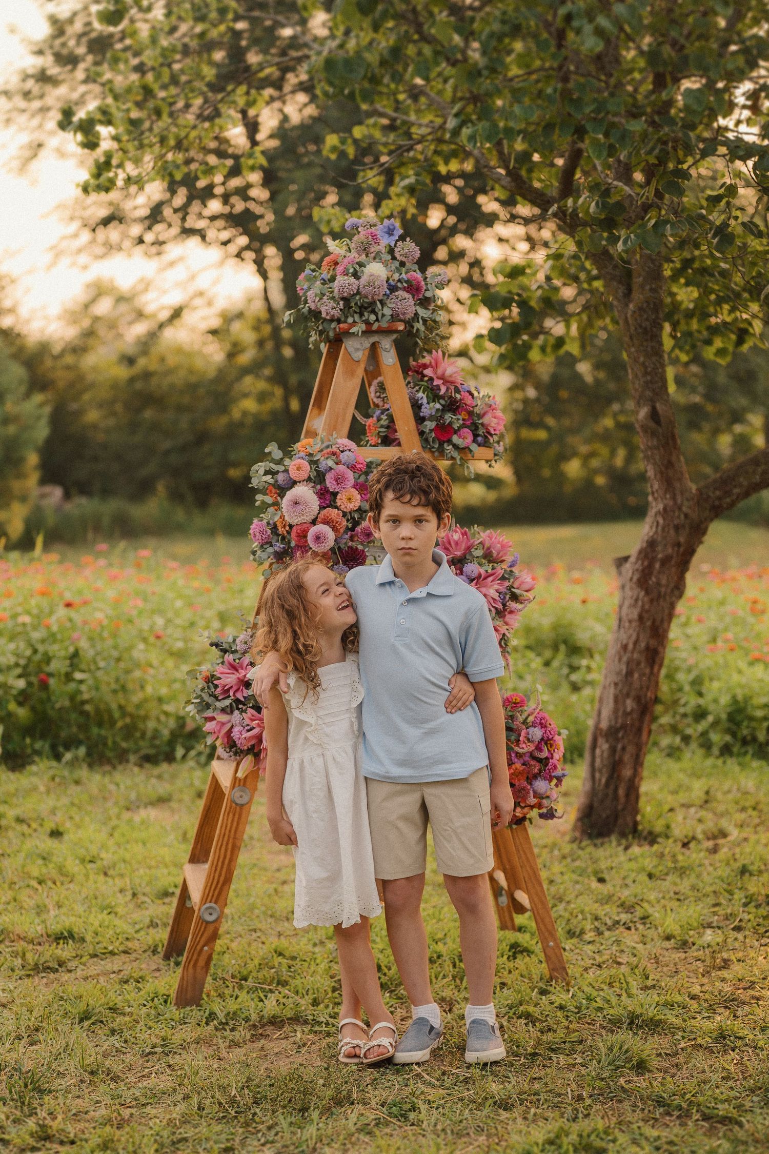 A couple poses near a wooden ladder decorated with pink and purple floral arrangements in a garden setting at sunset.