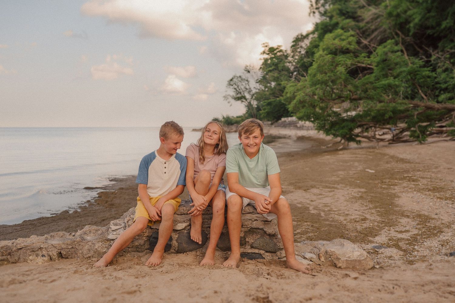 People relaxing on a sandy beach near water and trees during golden hour.