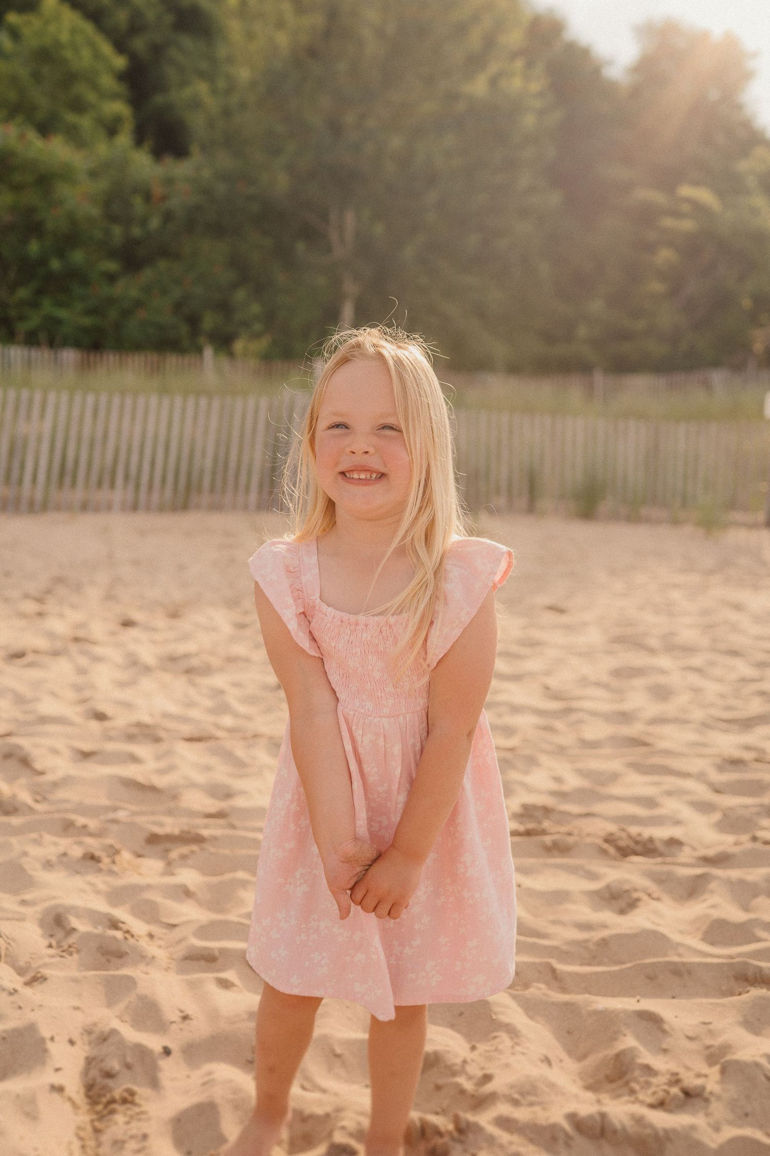 A child in a pink dress stands on a beach at sunset with wooden fencing and trees in the background.