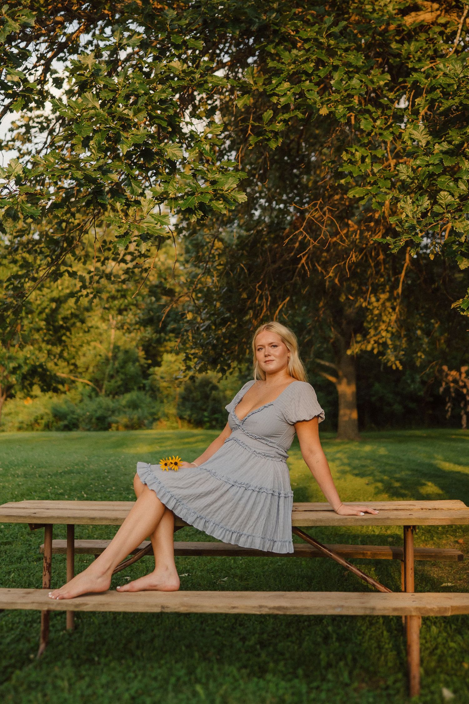 A person in a gray dress sits barefoot on a wooden picnic table surrounded by autumn trees at golden hour.