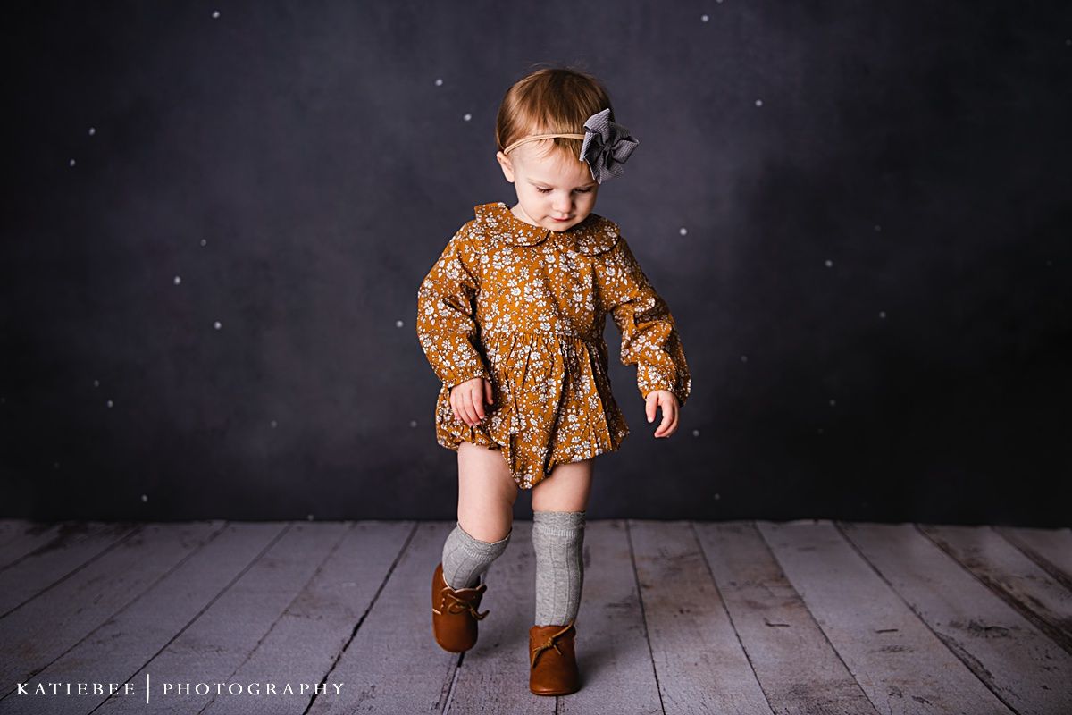 A child in a mustard floral dress and brown boots poses on wooden flooring.