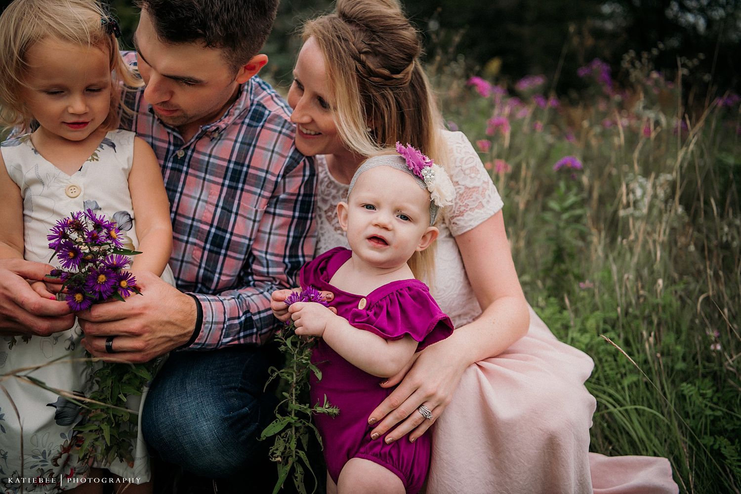 Family gathered closely together examining purple wildflowers in a summer meadow.