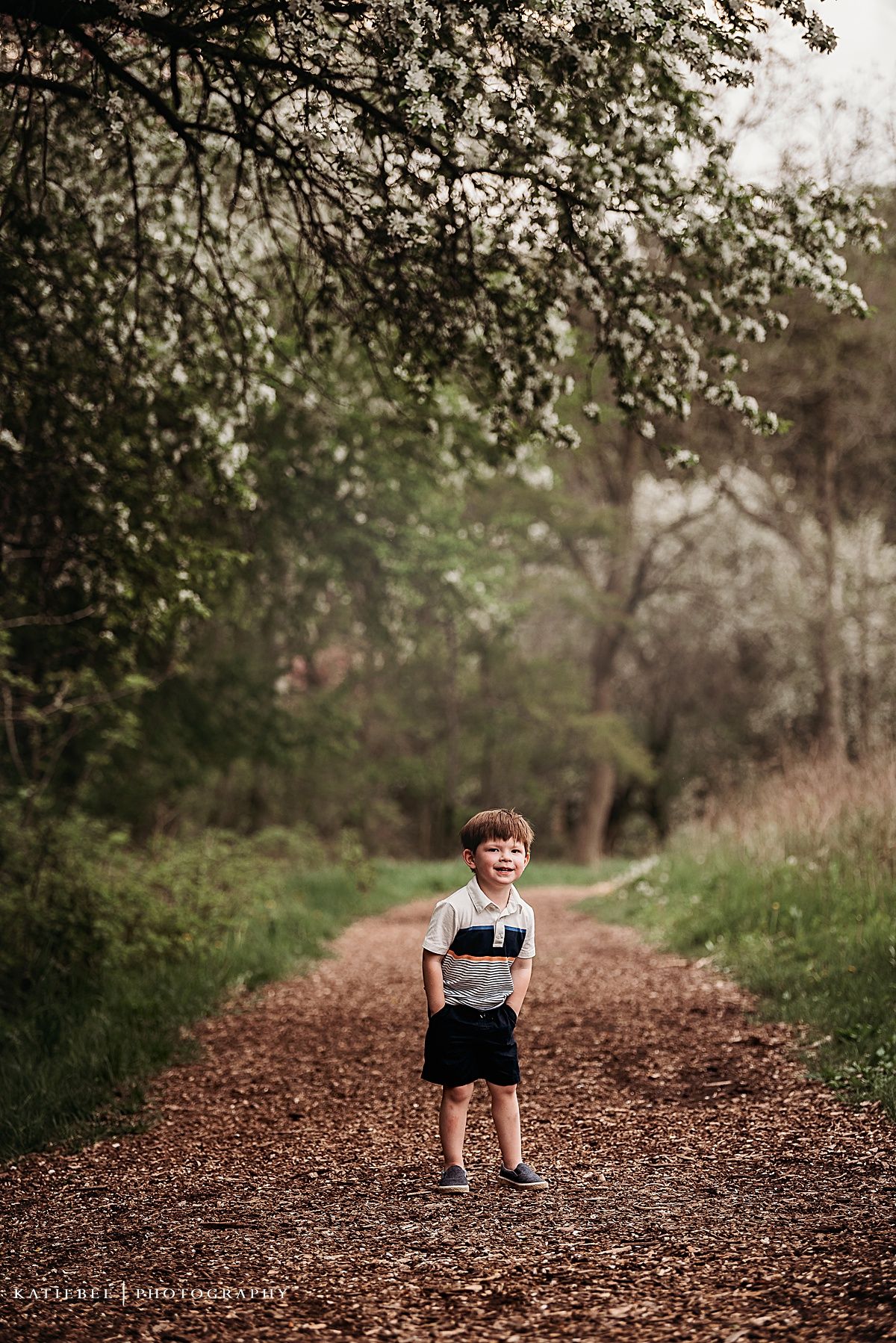 A child stands confidently on a tree-lined dirt path wearing a white and navy striped shirt.