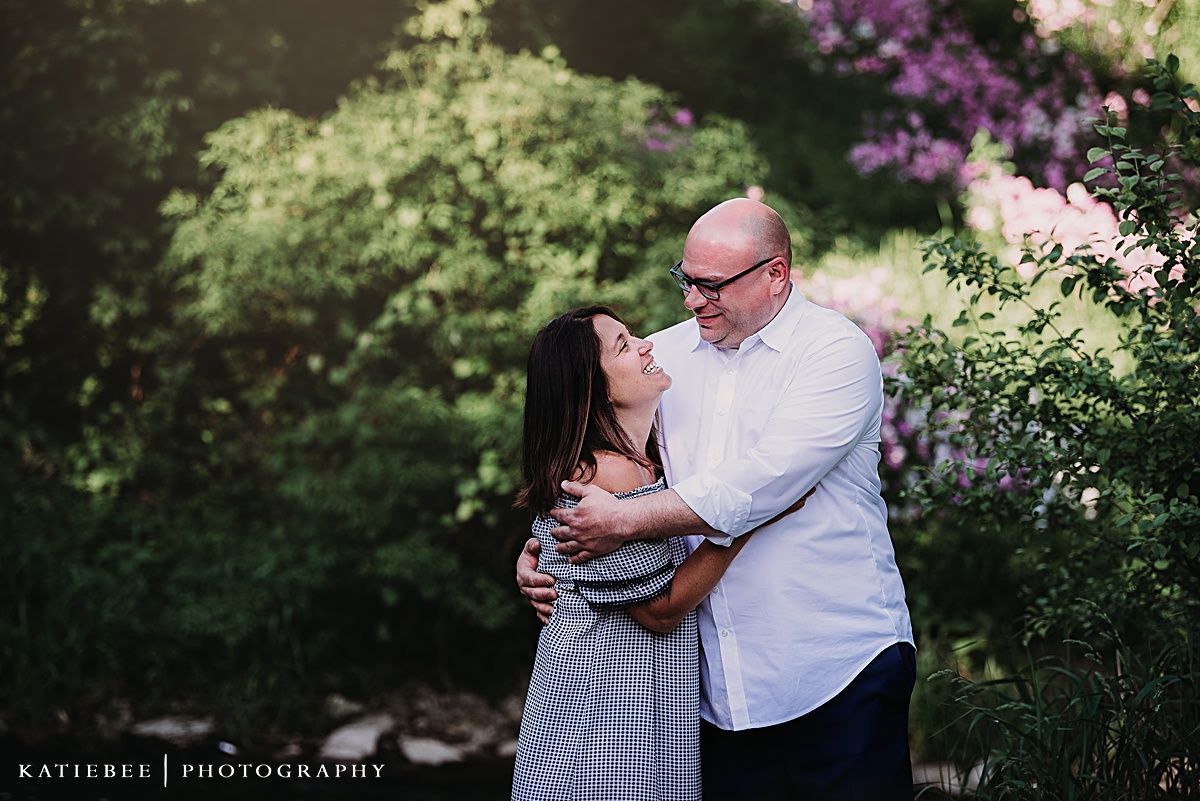 Intimate portrait of couple embracing in garden setting with purple flowers blooming in background.