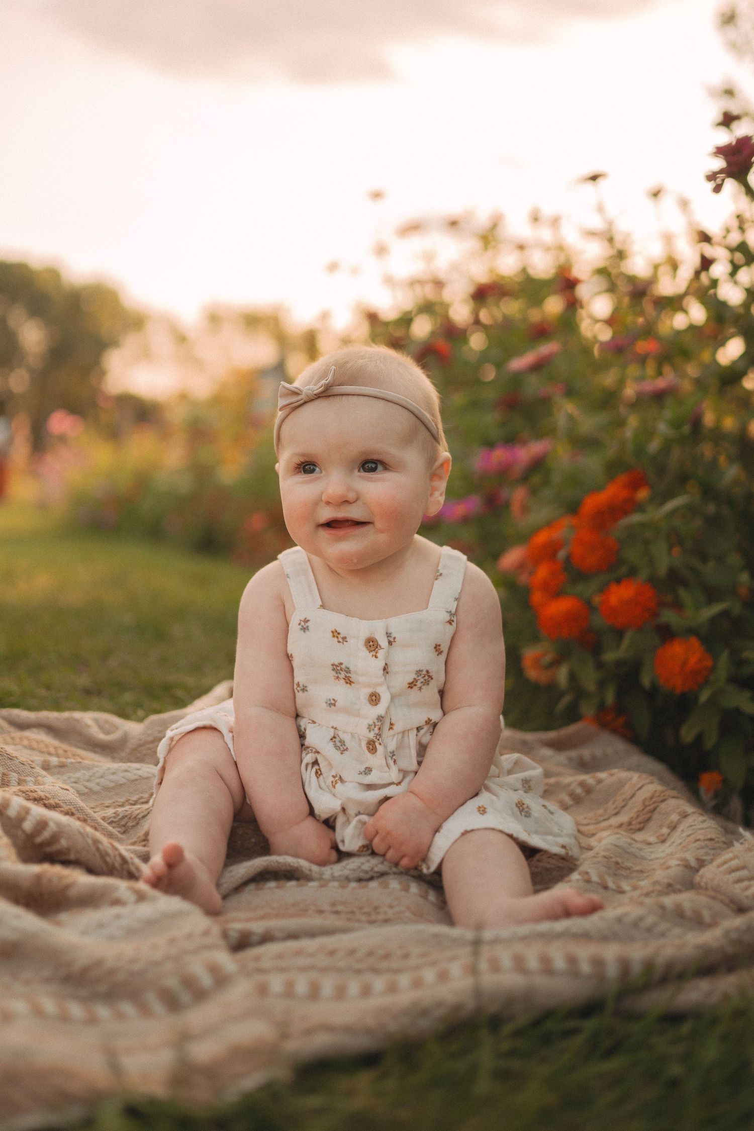 Baby in floral romper sits on textured blanket surrounded by orange and pink flowers.