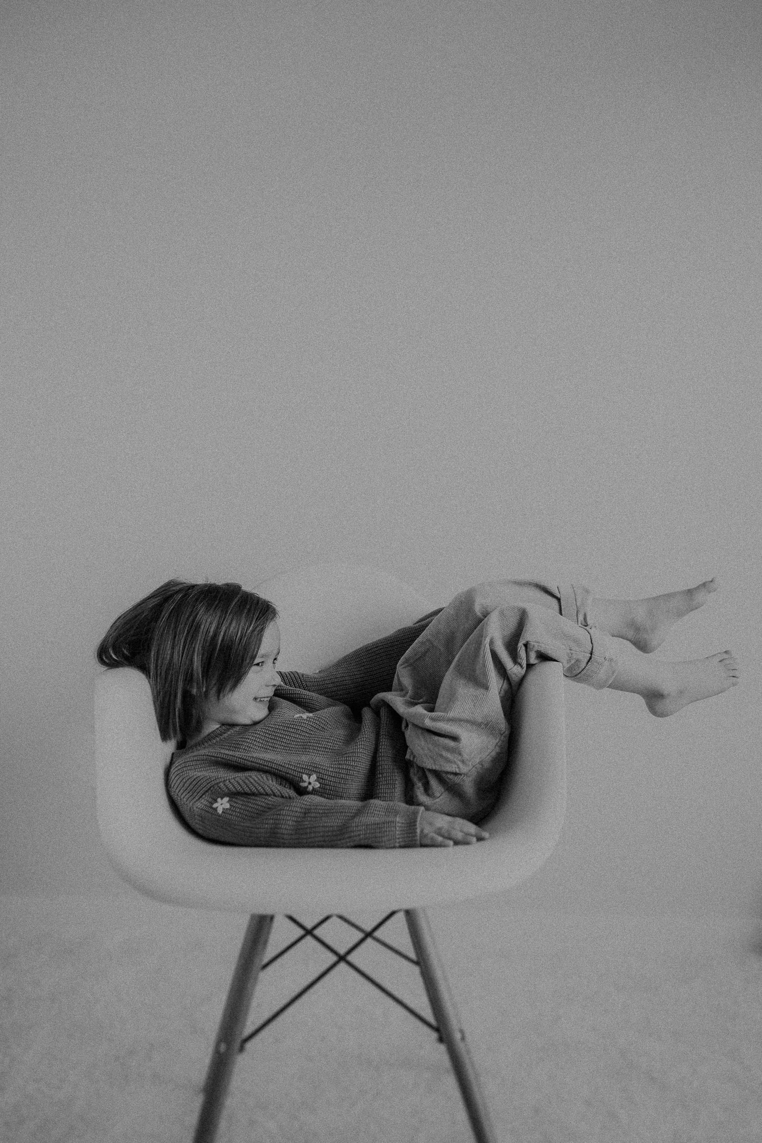 Child reclines sideways in modern chair during casual black and white photo session.
