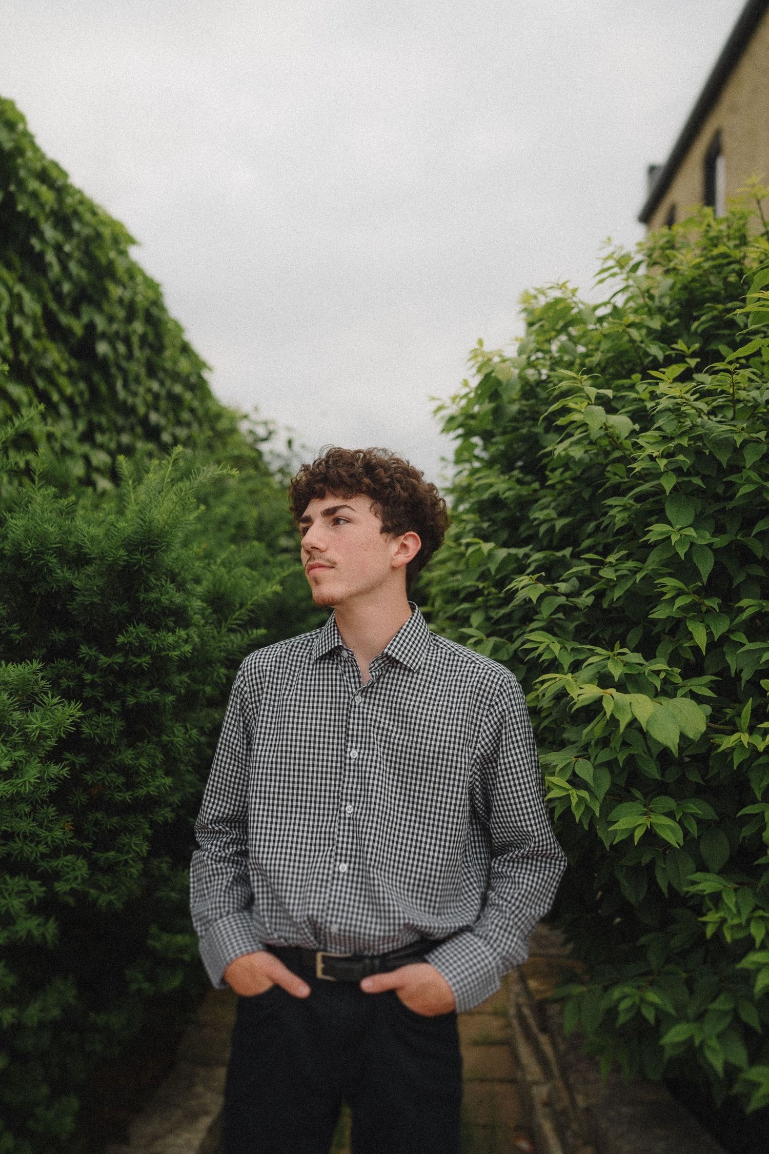 Person wearing a gingham check button-down shirt poses outdoors against green leafy backdrop on overcast day.