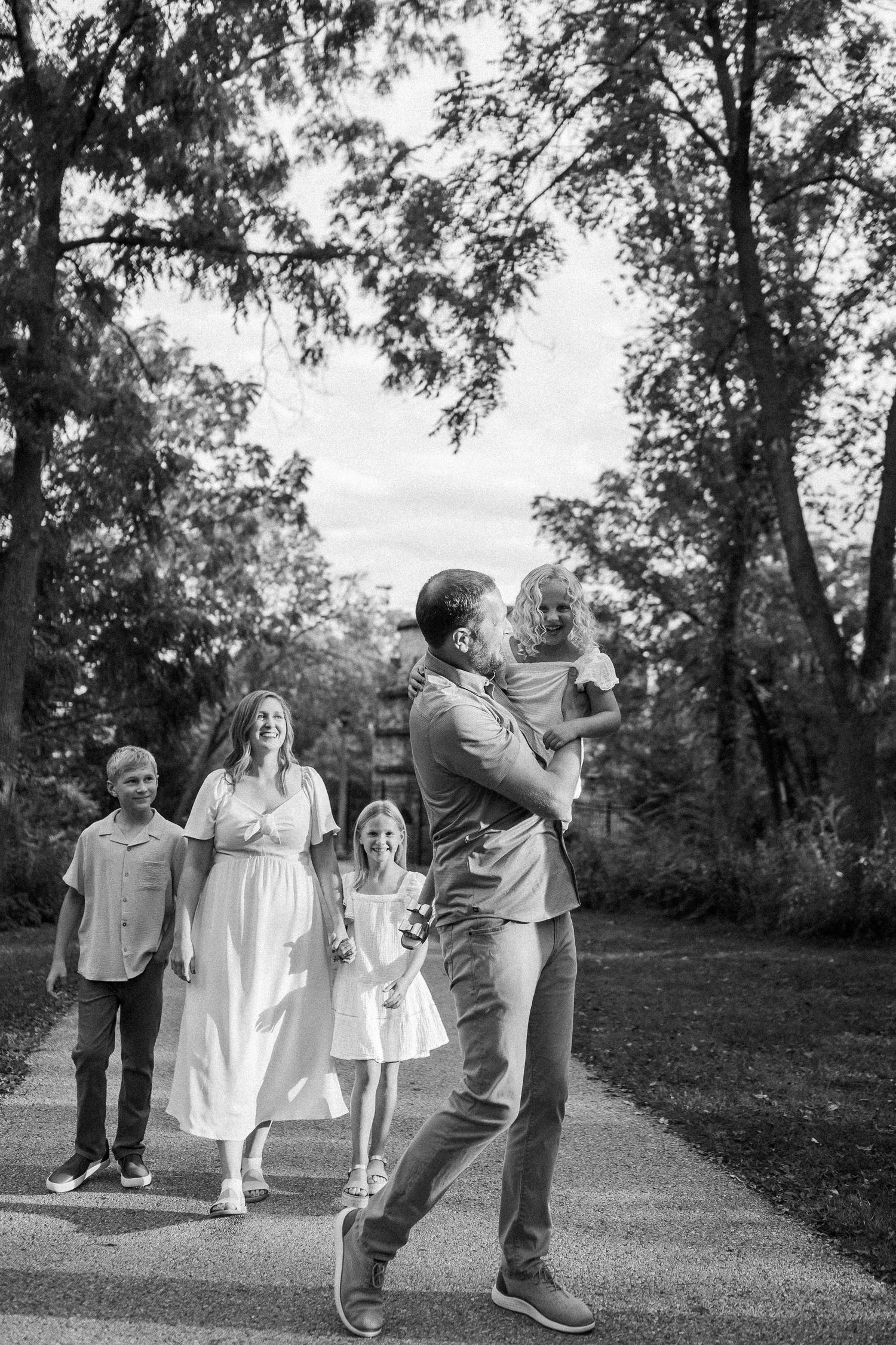 A group in white and casual attire plays and interacts joyfully on a grassy lawn with trees in the background.