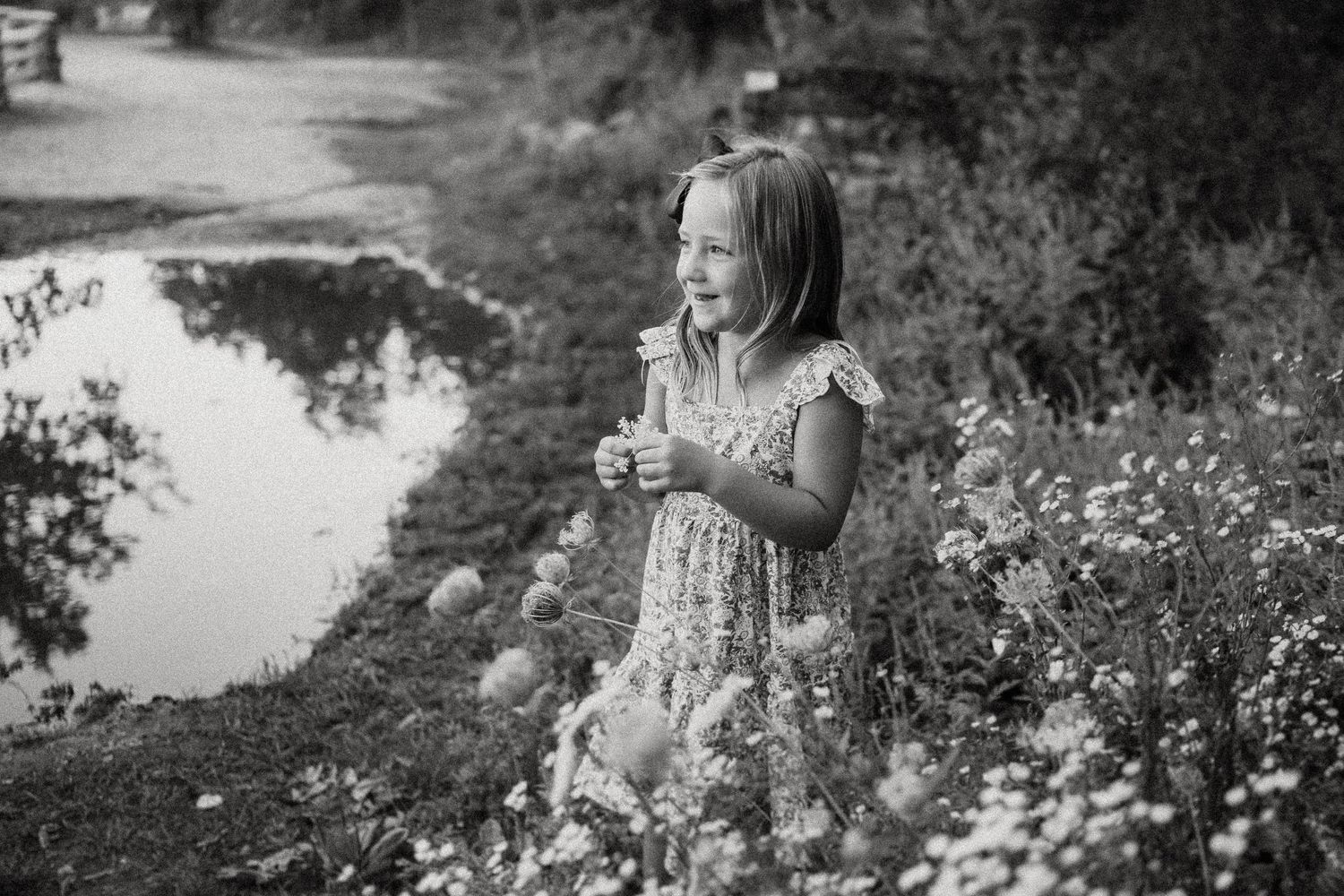 Black and white portrait of a child standing among wildflowers by a pond.