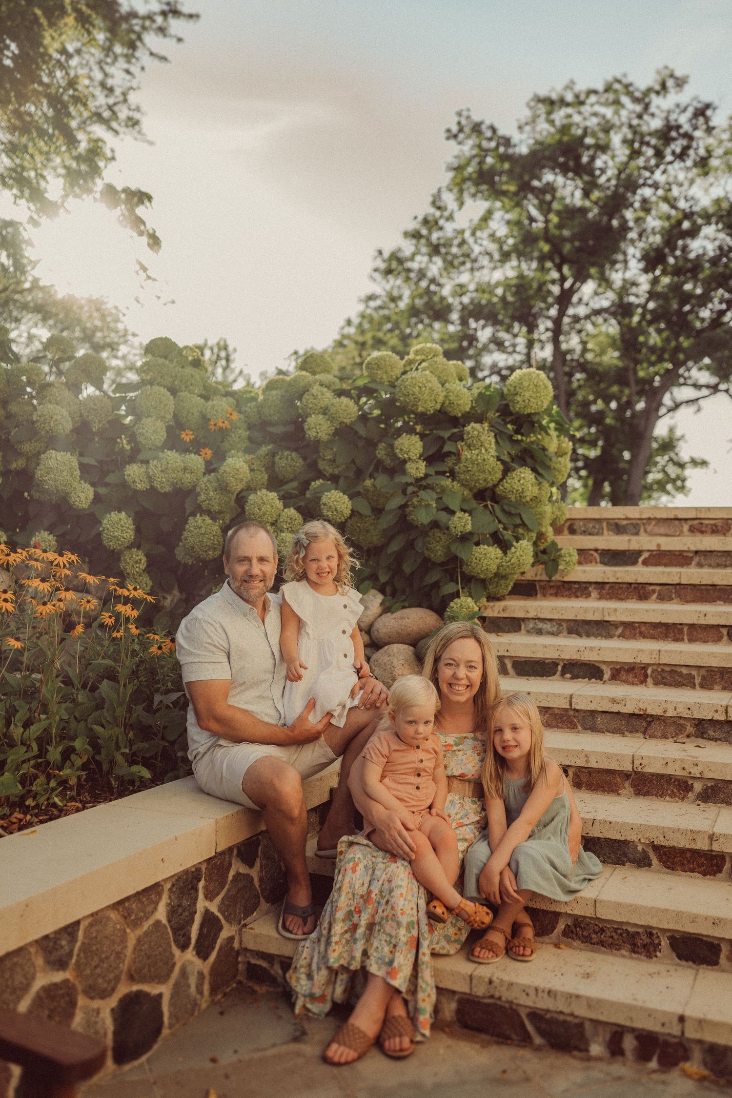 Family portrait on stone garden steps with lush hydrangea bushes and warm sunset lighting.