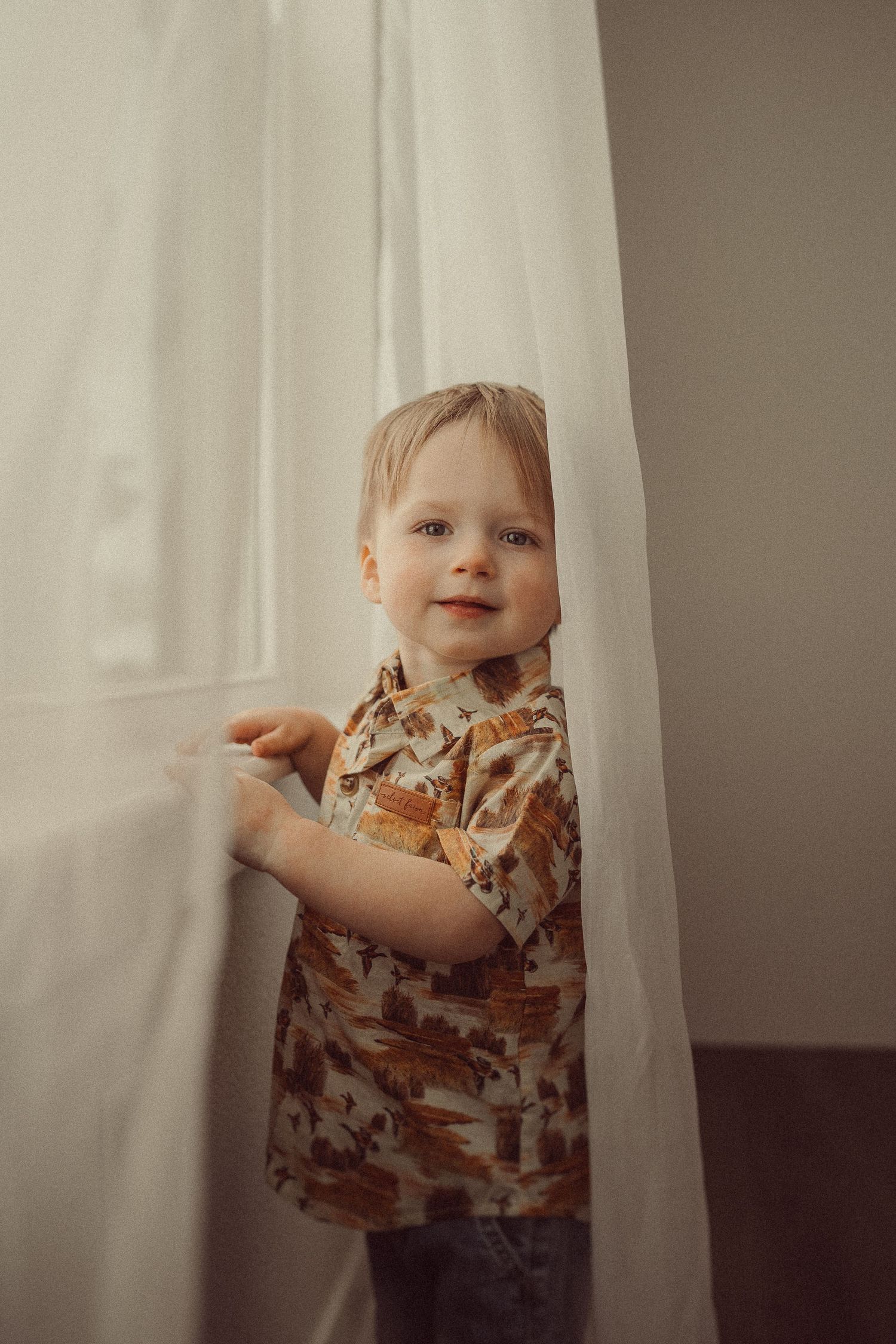 Child in brown patterned shirt peeking from behind white curtain.