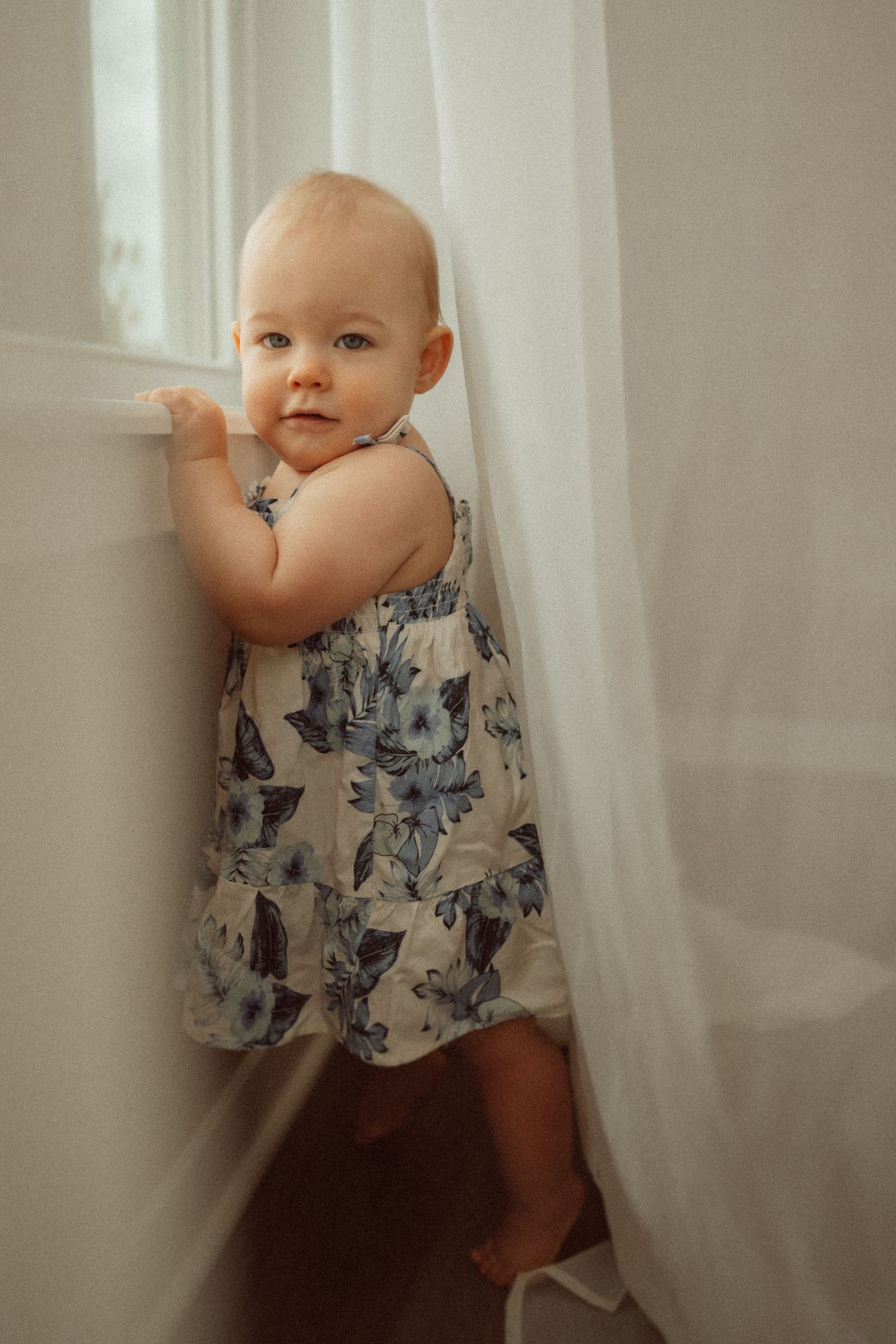 Baby in patterned dress stands beside window holding white curtain.