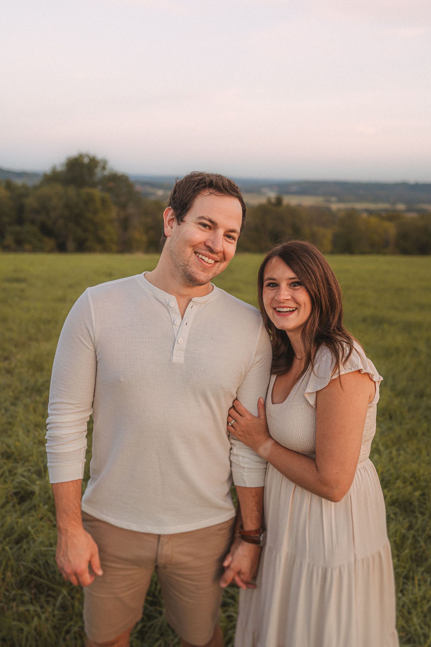 Couple poses together in a grassy field during golden hour with rolling hills in the background.