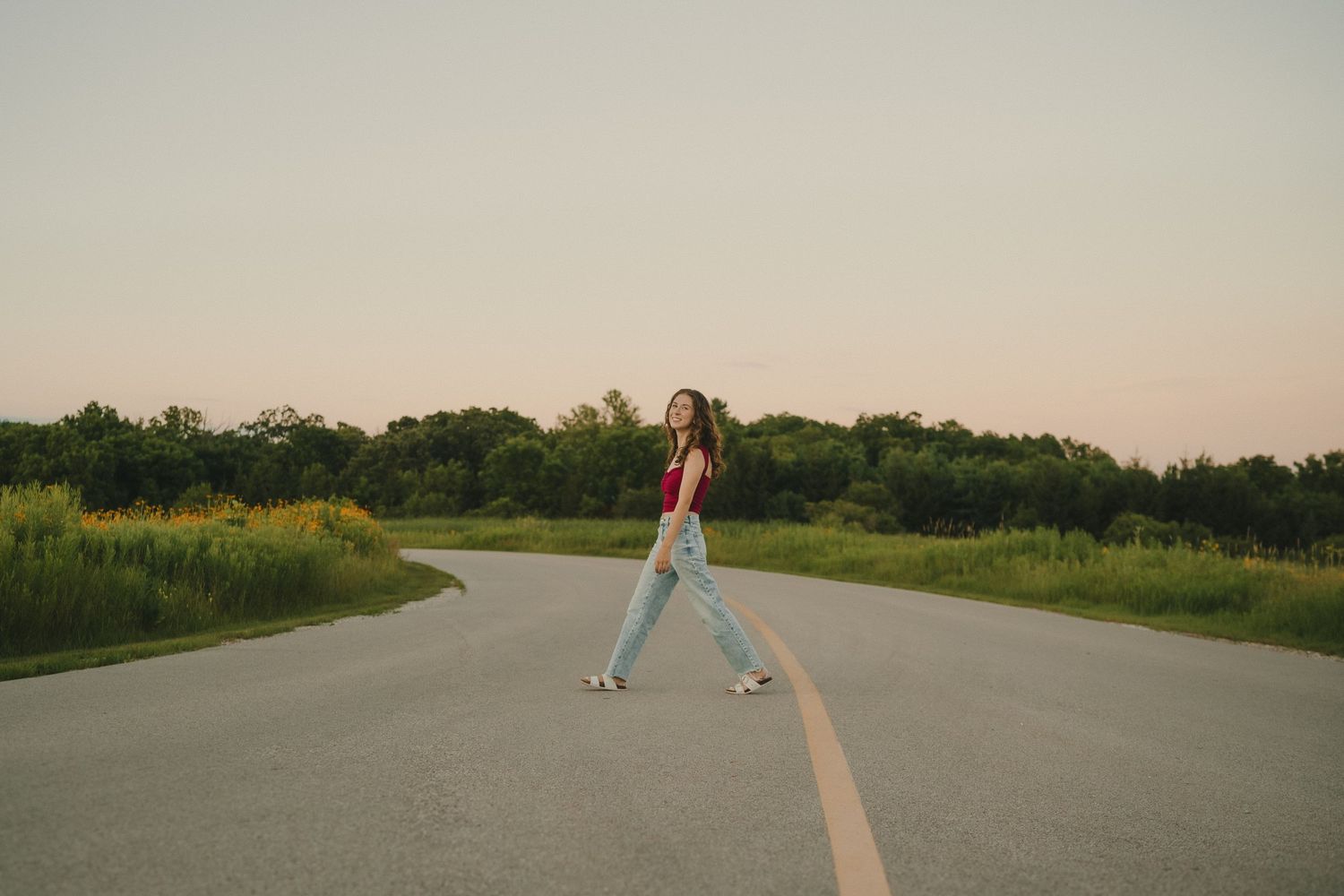Someone in jeans walking down the middle of an empty curved road at sunset with wildflowers along the sides.