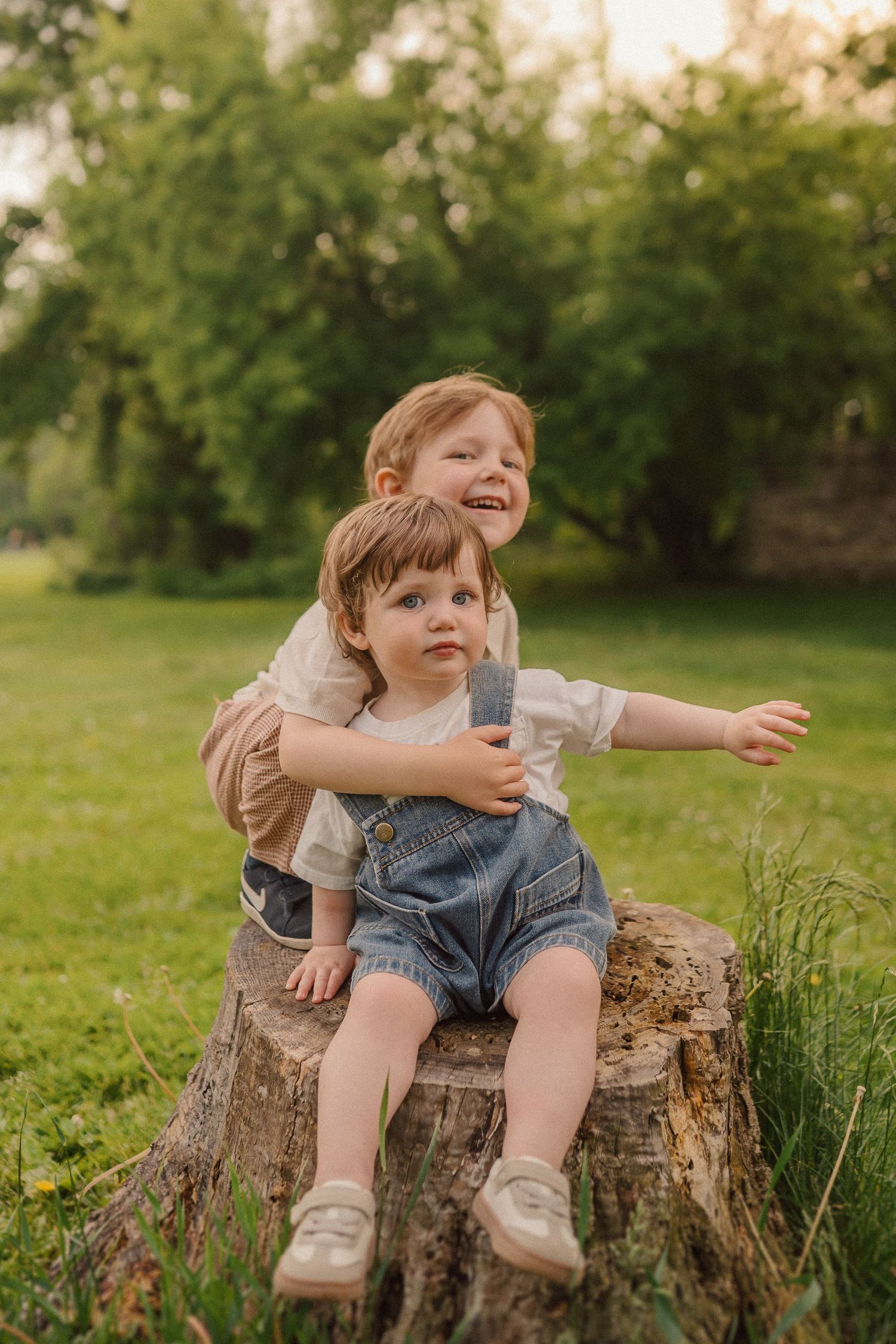 Two children share a joyful moment while sitting on a wooden tree stump in a sunlit garden.