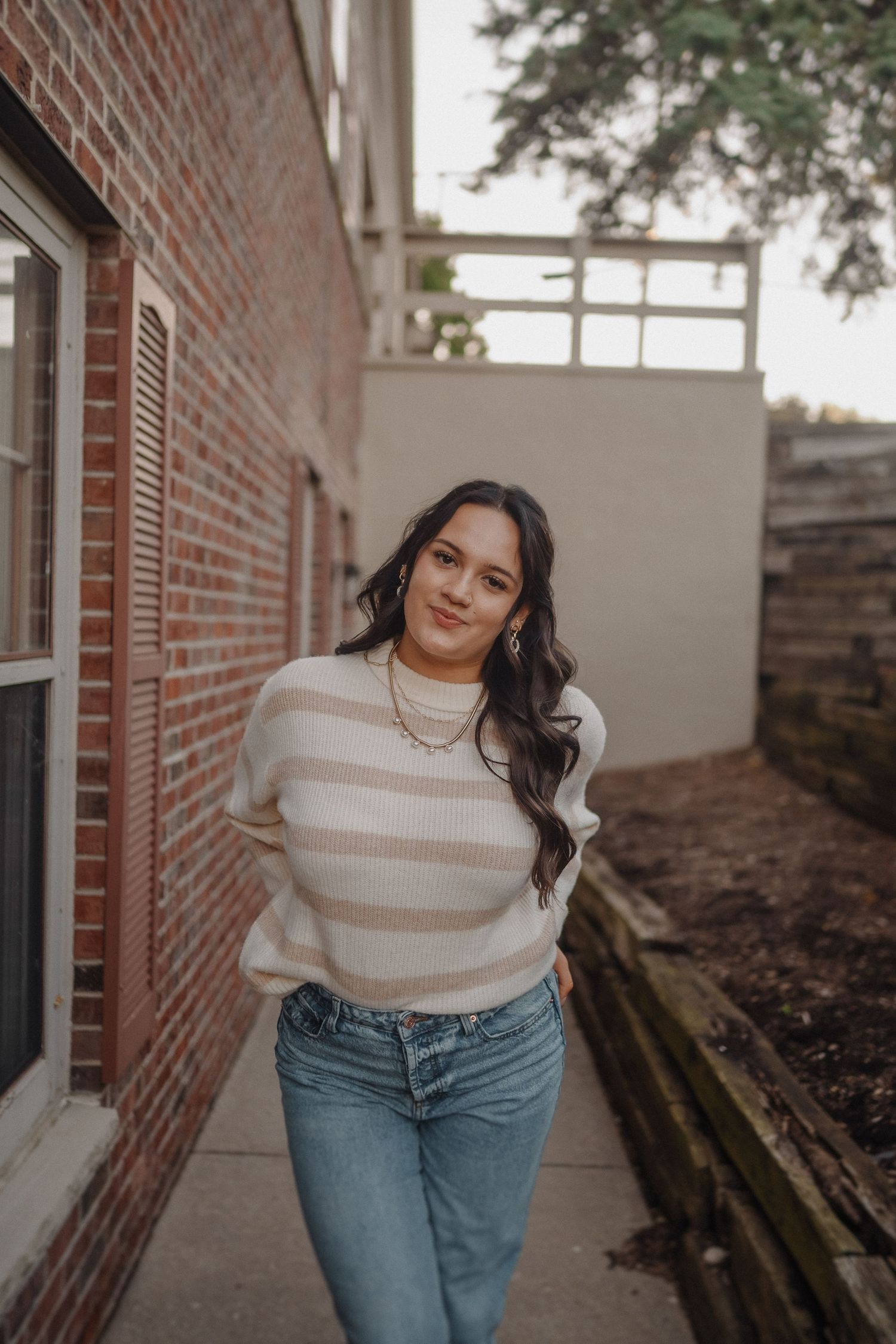 A person in a striped sweater and jeans stands casually against a brick wall background.