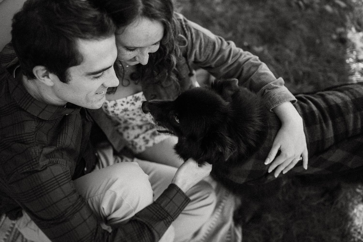 A black and white intimate moment showing a couple petting their dog outside.