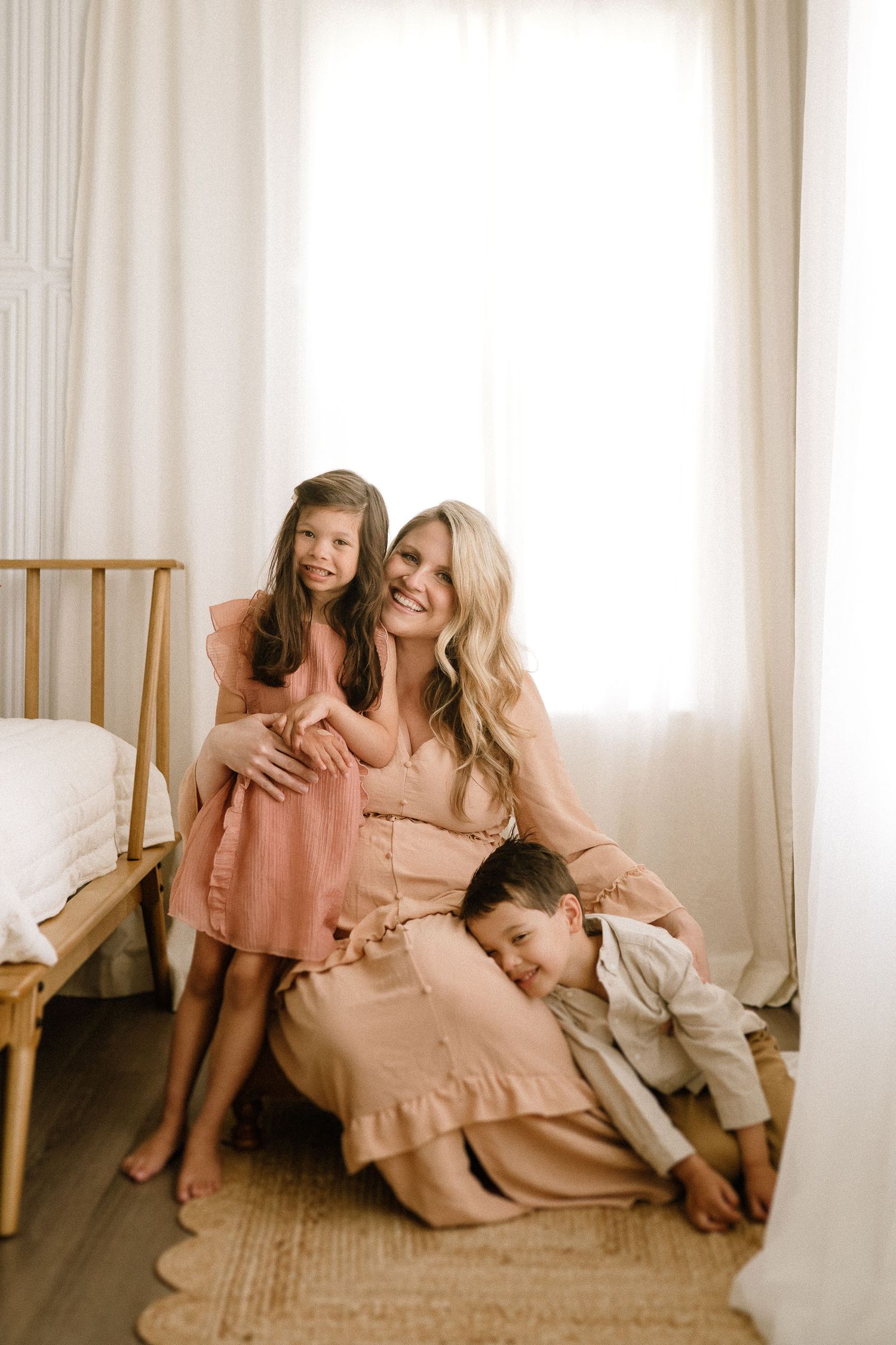 Sweet family bonding scene with mother and children in matching neutral tones sitting on woven rug by window.