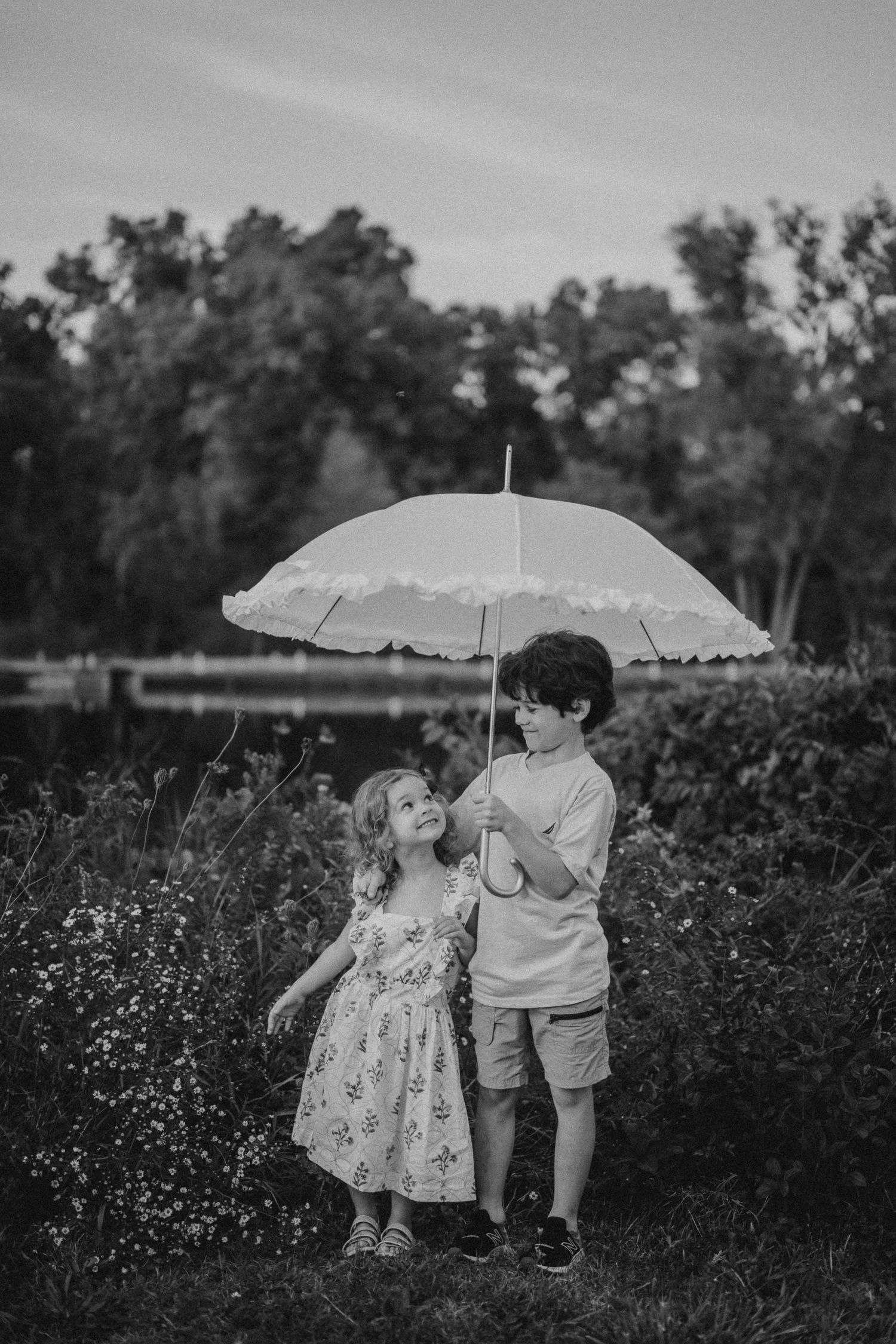 Black and white photo of two children sharing an umbrella in a meadow.