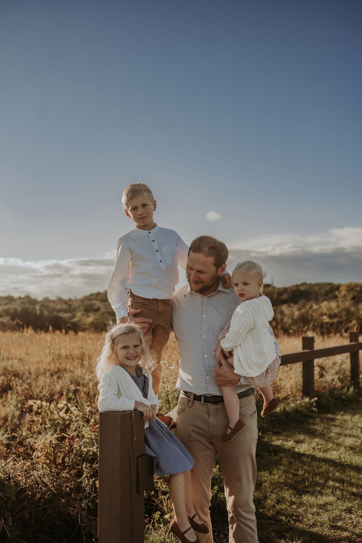 A warm sunset family portrait with everyone gathered around a wooden fence post in a rural setting.