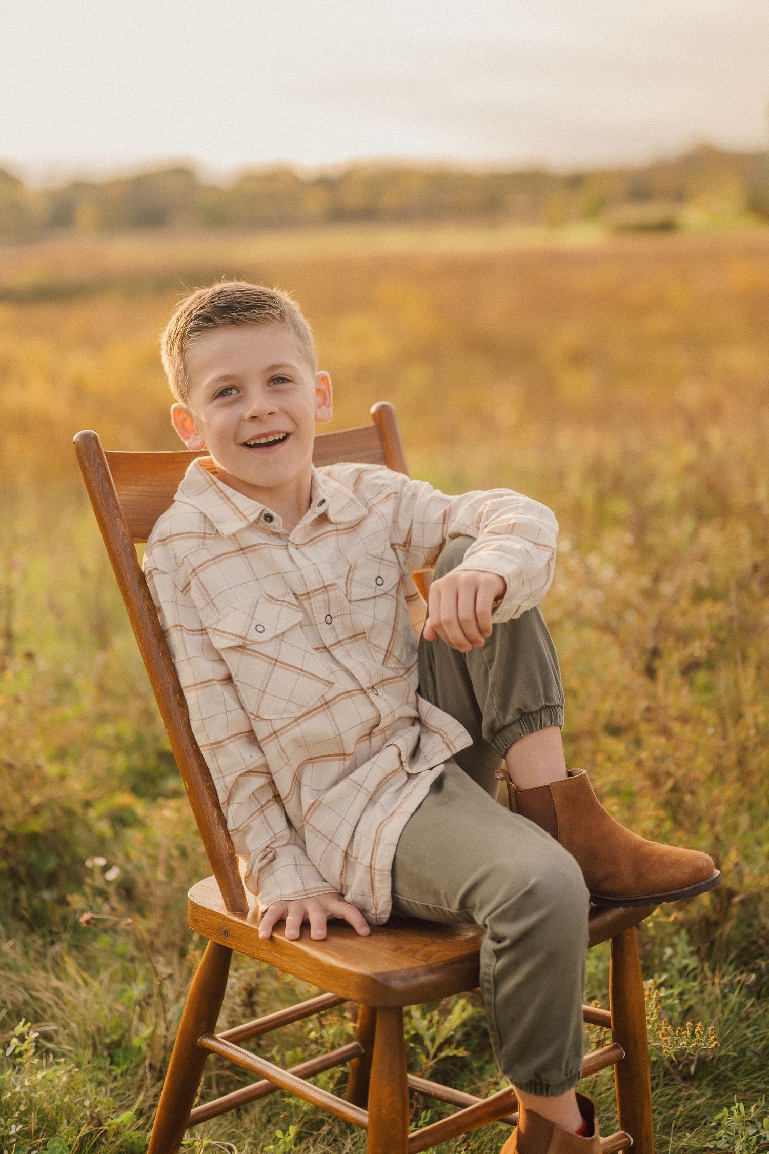 Child relaxing on wooden chair in golden sunlit field wearing plaid shirt and casual attire.