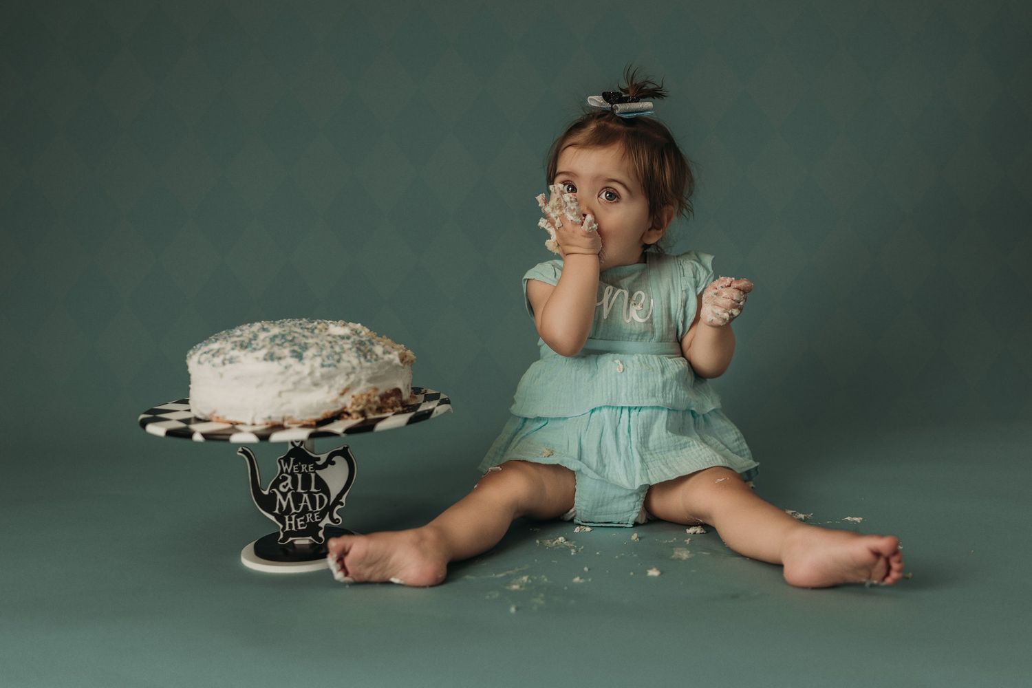 A small figure in a mint green dress sits beside a white cake on an ornate cake stand against a dark green backdrop.