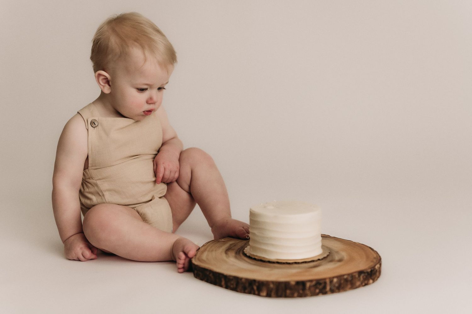 Baby in tan outfit examines white cake displayed on rustic wood slice against beige background.