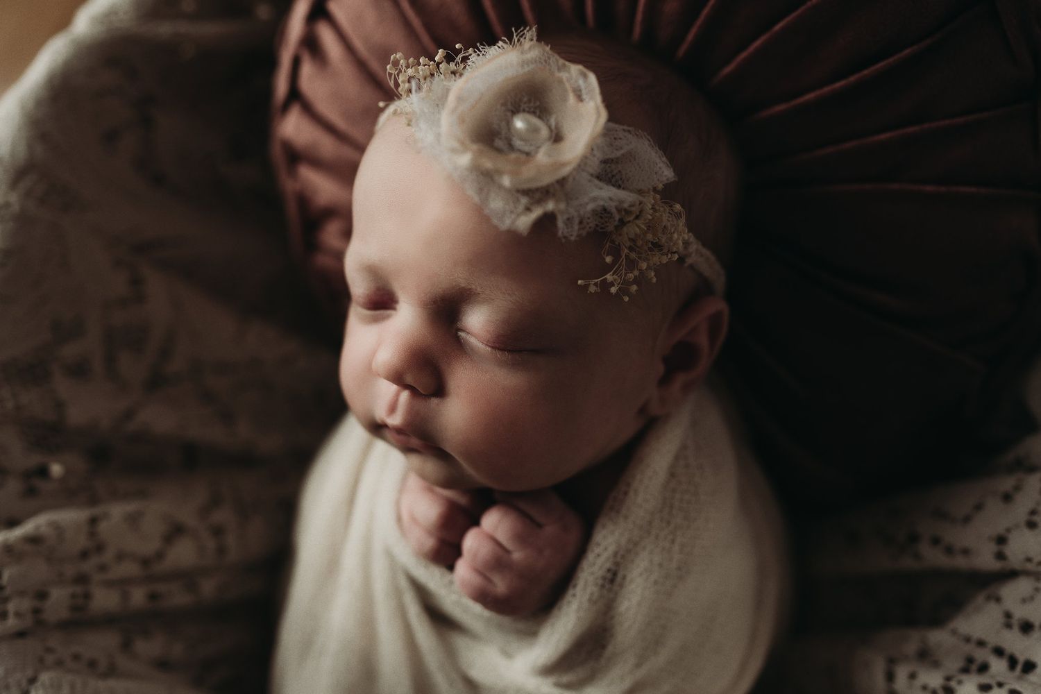 Newborn sleeping peacefully in soft light wearing delicate headpiece and wrapped in cream colored swaddle.