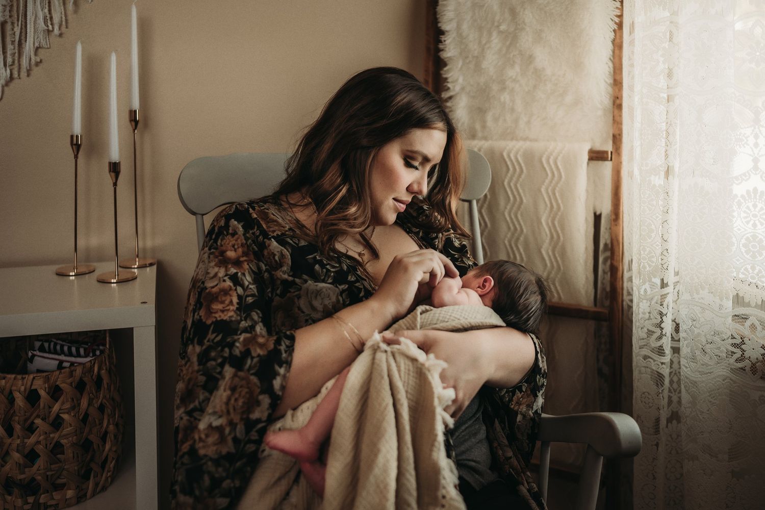 Tender nursing moment in a rocking chair beside white curtains and decorative candles.