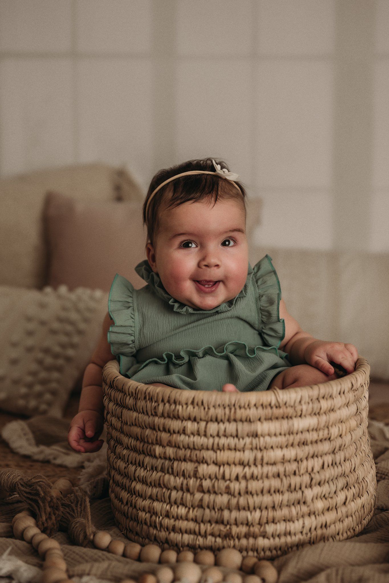 A baby in a mint green dress sits happily in a woven basket on textured blankets.