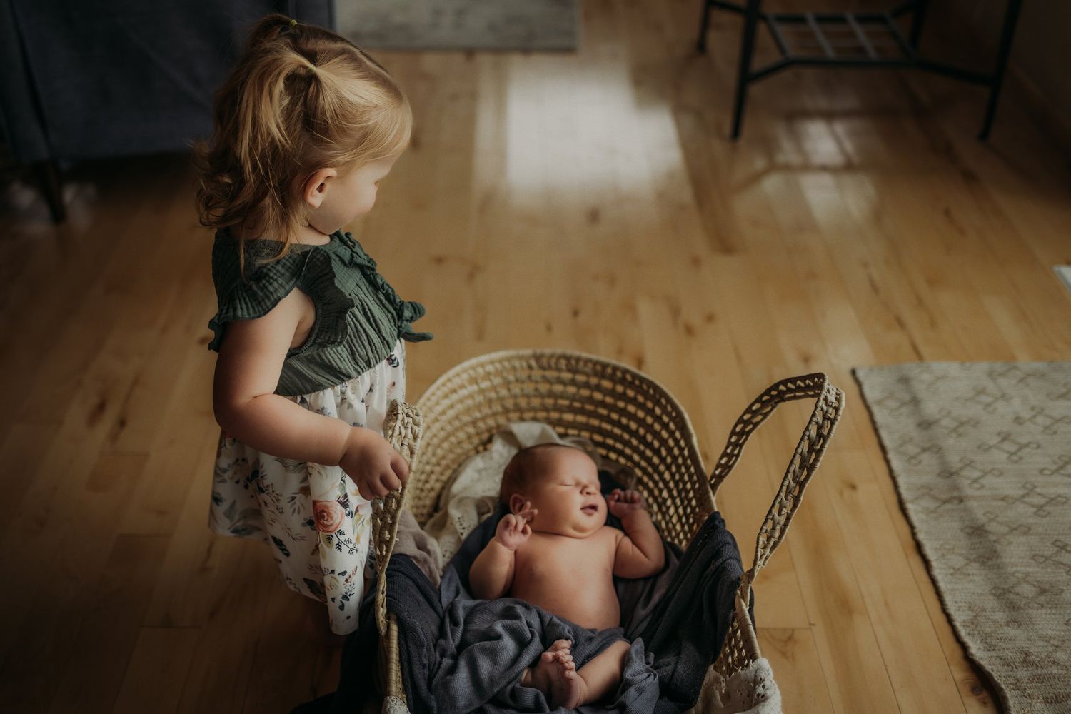 Young child peeks into wicker bassinet containing newborn sibling.