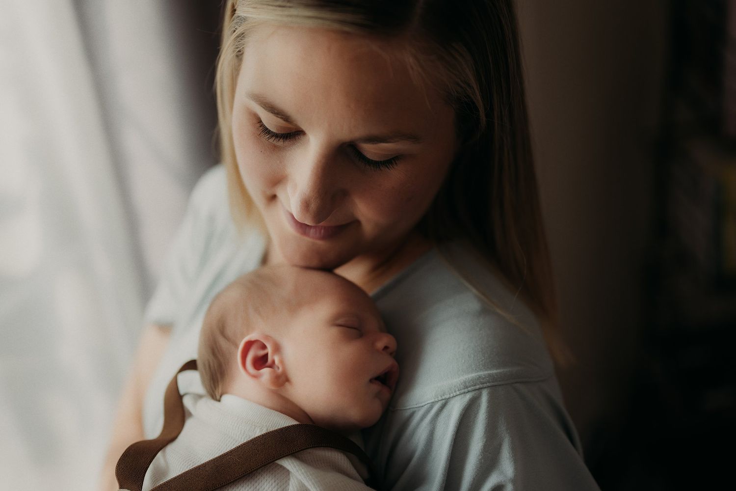 Parent holds sleeping newborn baby close while standing in soft window light.