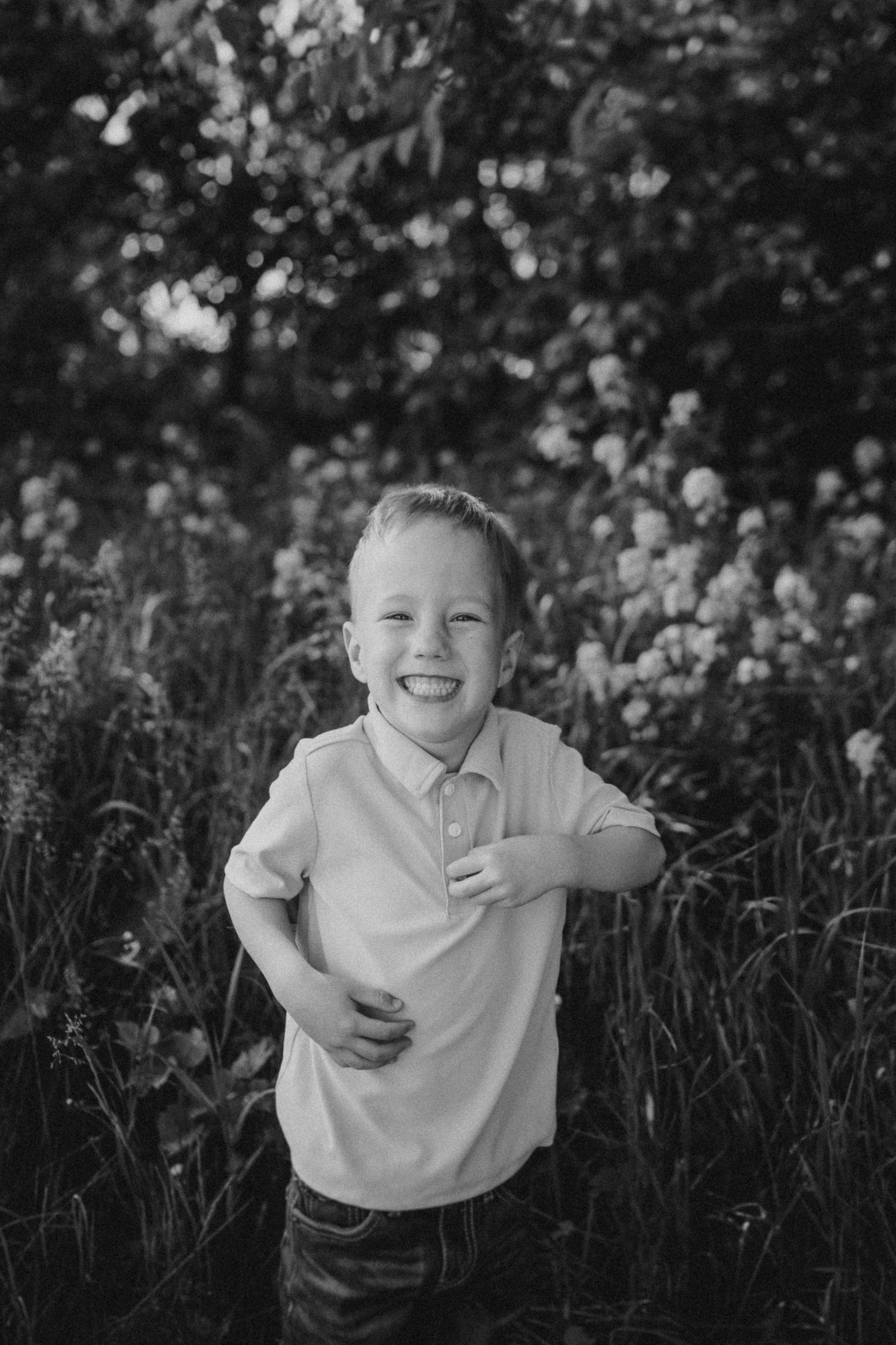A black and white portrait shows a laughing child in a polo shirt among wildflowers.