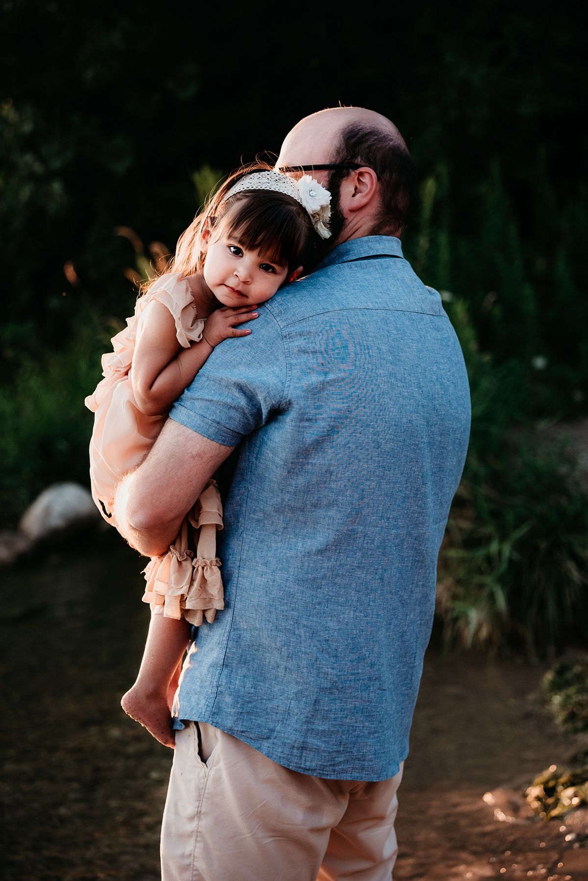 Child in pink dress rests head on shoulder against natural outdoor setting.