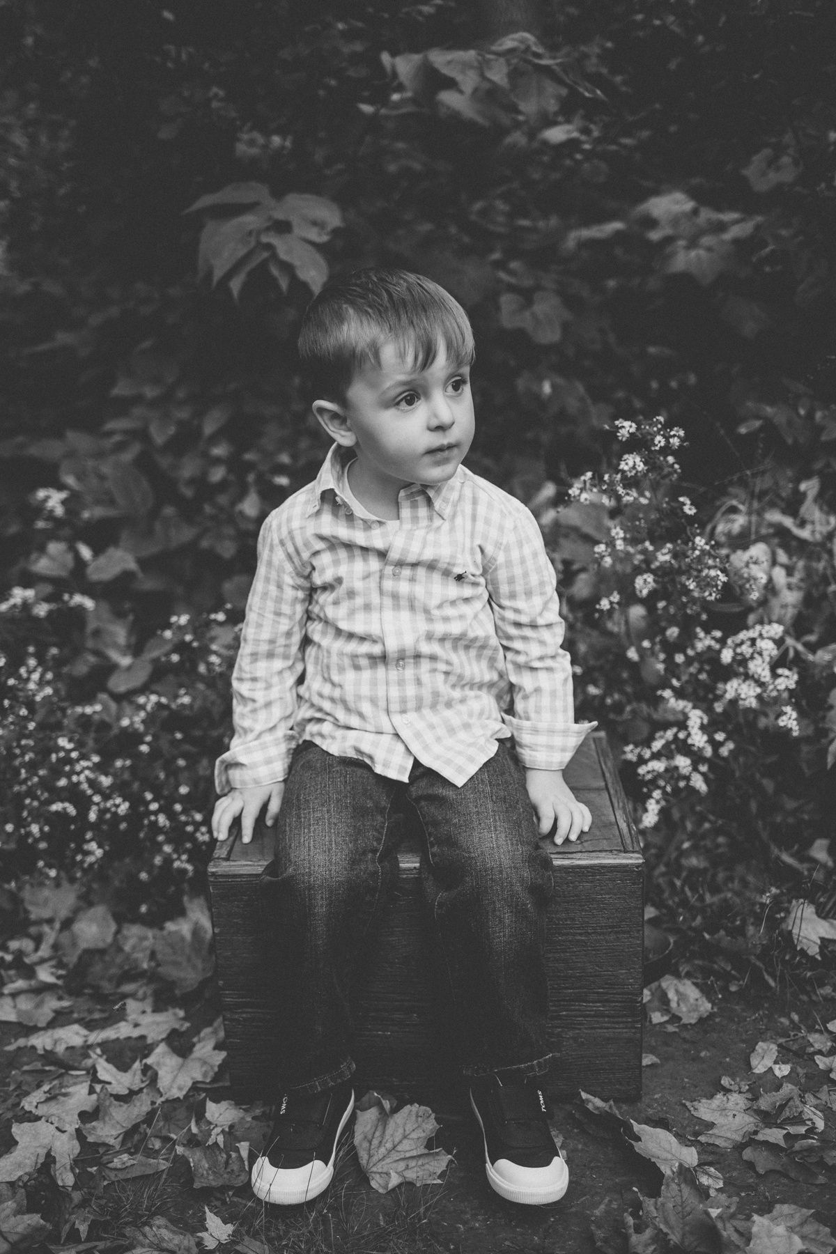Black and white portrait of young boy in checkered shirt sitting on wooden crate outdoors.