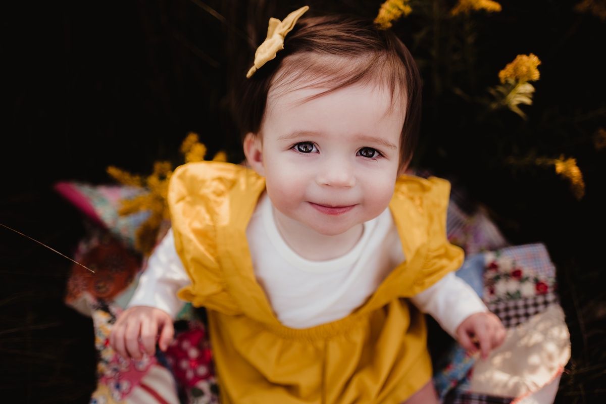 Close-up portrait in mustard yellow dress with butterfly hair clip against dark foliage.