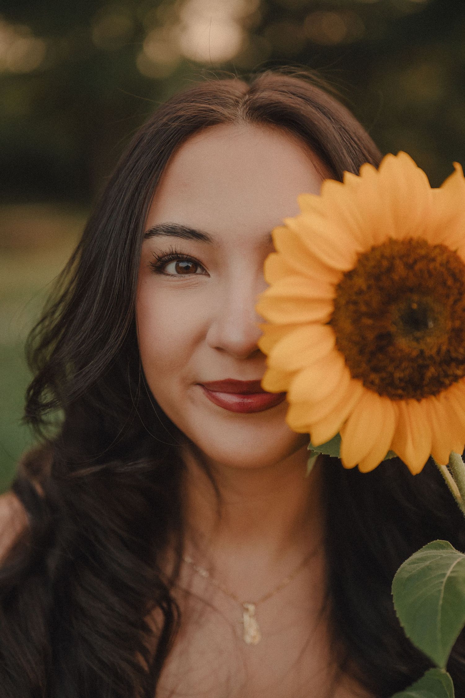 Close-up portrait of a young woman with dark wavy hair holding a bright yellow sunflower over half her face outdoors.