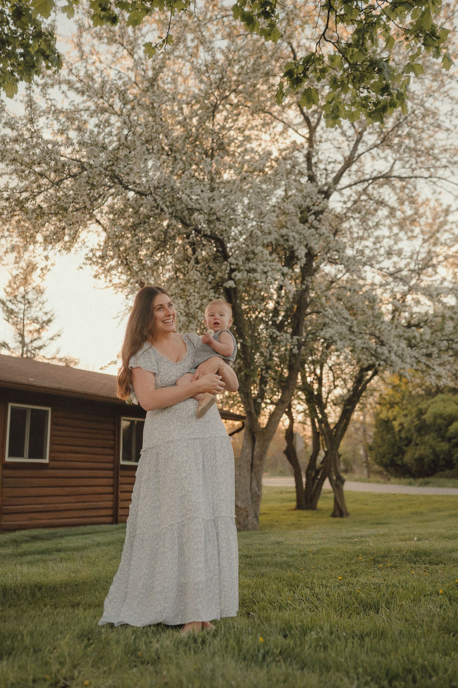 Smiling mother in white floral maxi dress holding laughing baby near blooming white tree at golden hour sunset.