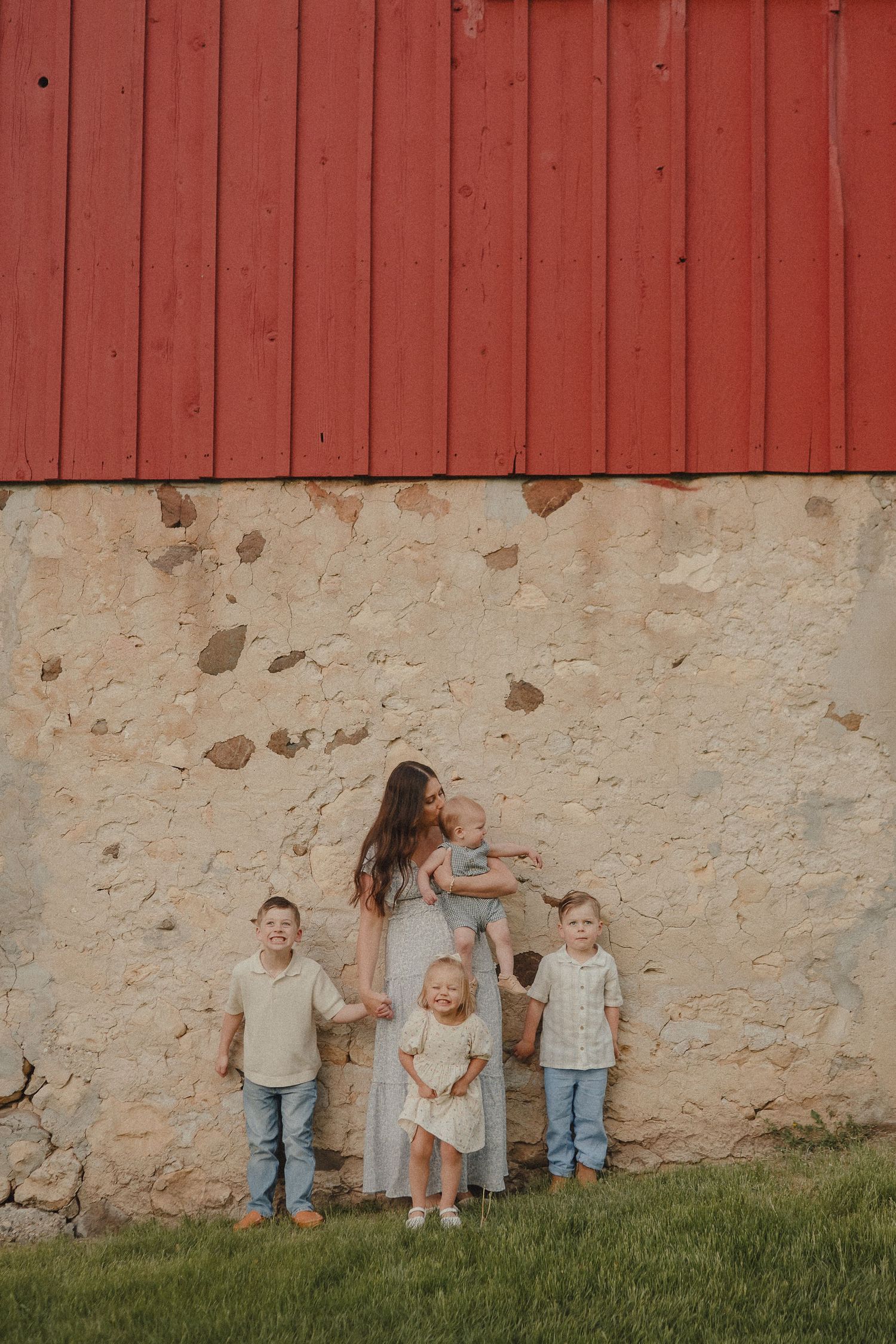 Mother with four young children posing against a rustic stone and red barn wall outdoors.