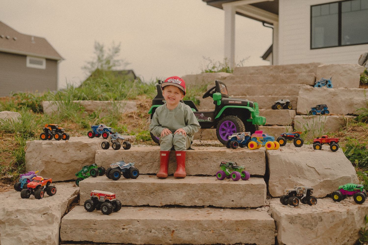 Toddler boy sitting on stone steps smiling, surrounded by collection of colorful monster truck toys outdoors.