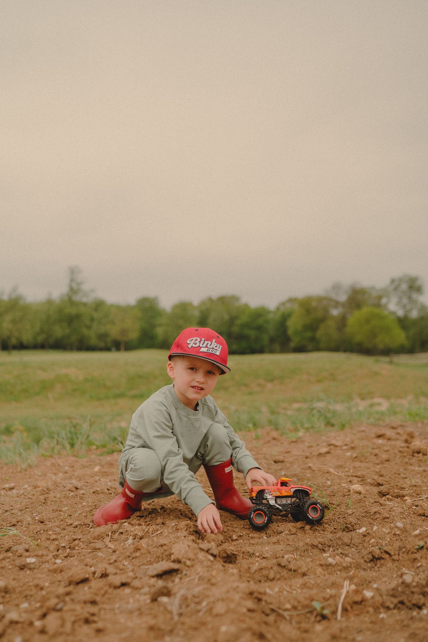 Young boy in red cap and boots plays with toy monster truck in a dirt field.